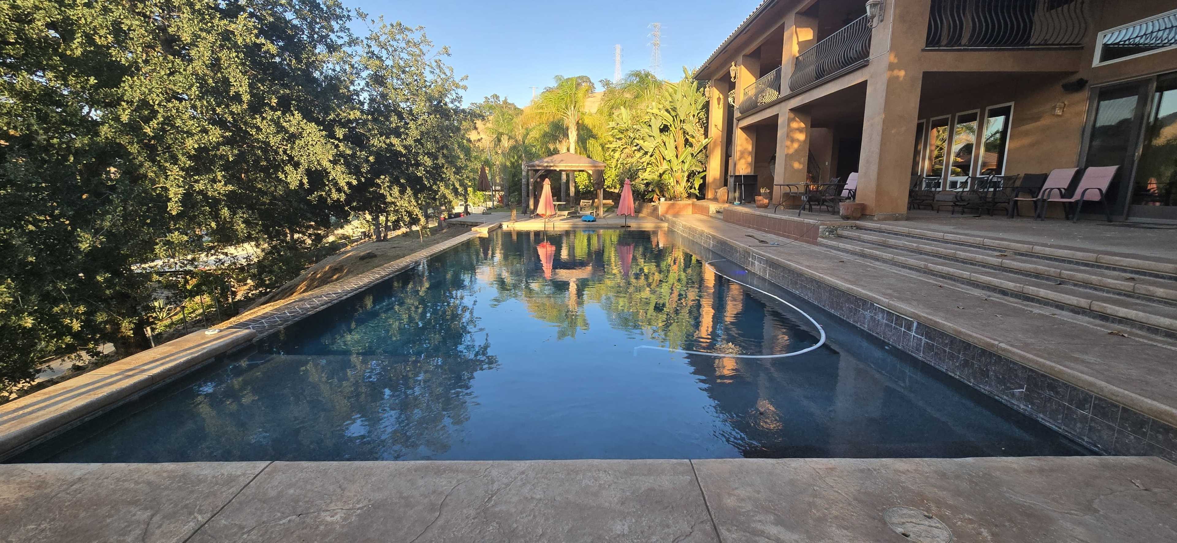The image shows a rectangular swimming pool surrounded by tropical vegetation and a two-story house, with patio furniture arranged beside the pool.