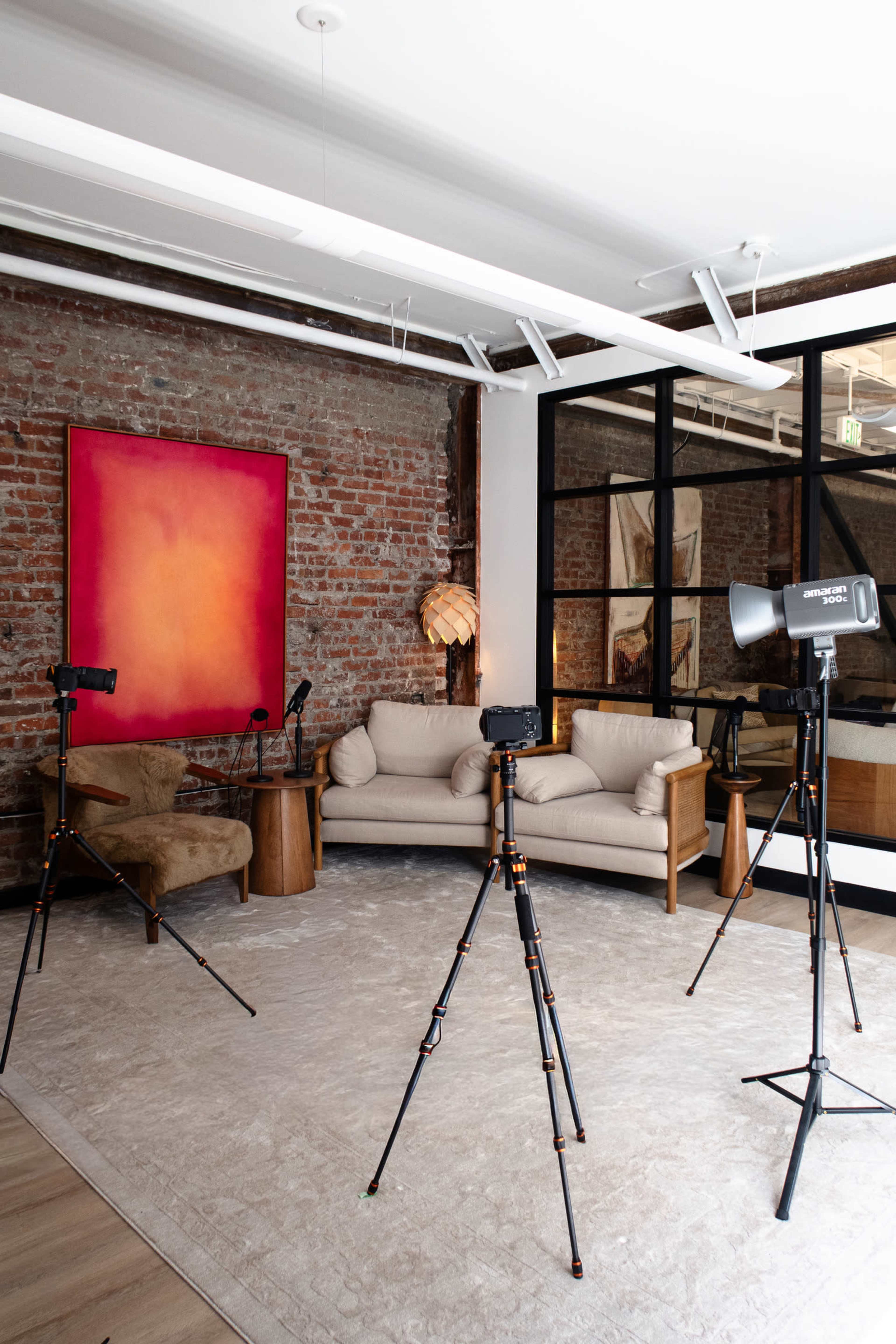 The image shows a bright, modern interior space featuring a sofa, a side table, and multiple cameras on tripods, with a large abstract painting on the wall.