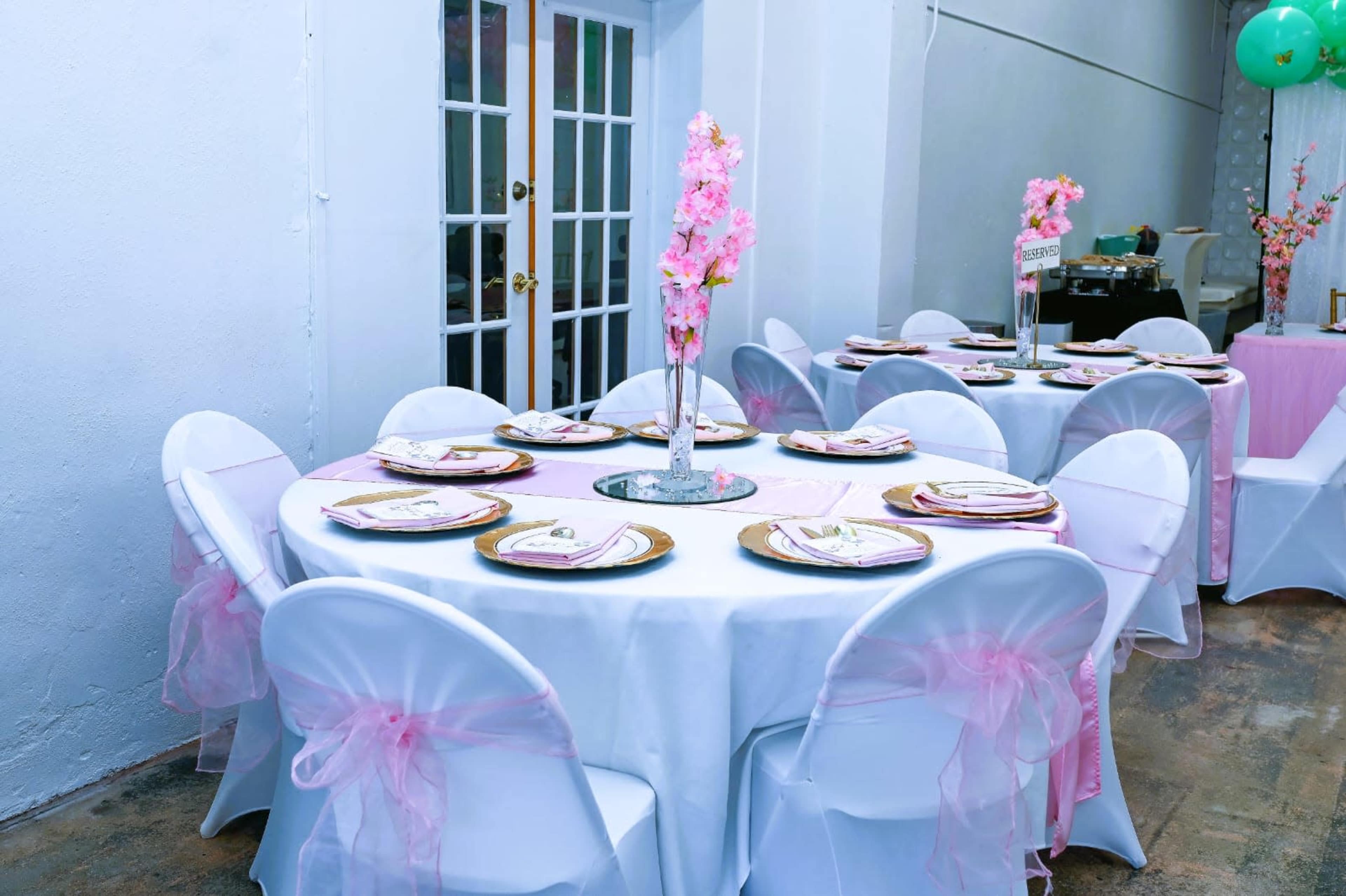 The image shows a banquet setup with round tables covered in white tablecloths, each adorned with pink decorations and centerpieces featuring floral arrangements.