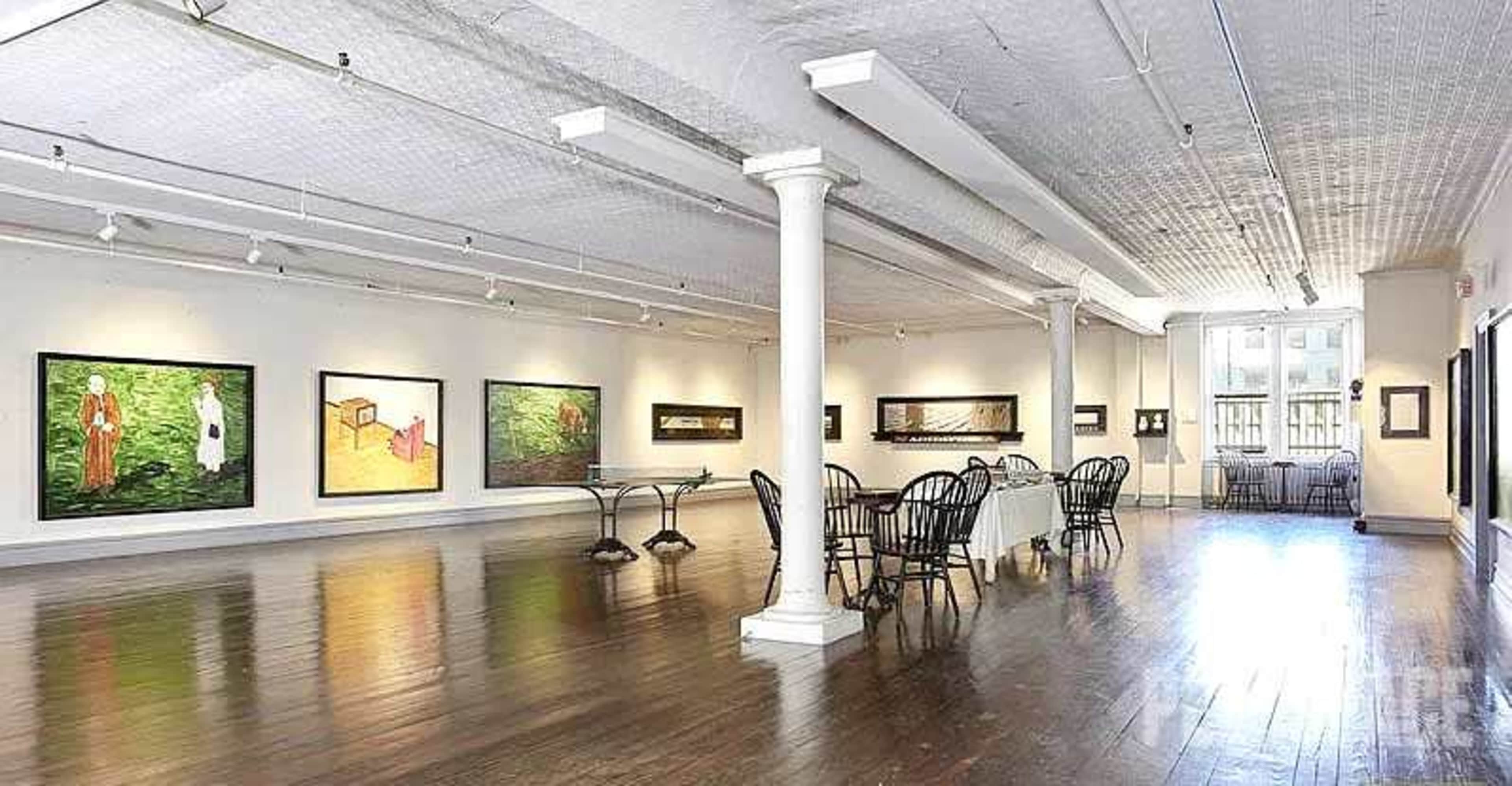 The image shows an art gallery with several framed paintings on the walls and a dining table set with chairs in the center of the room.