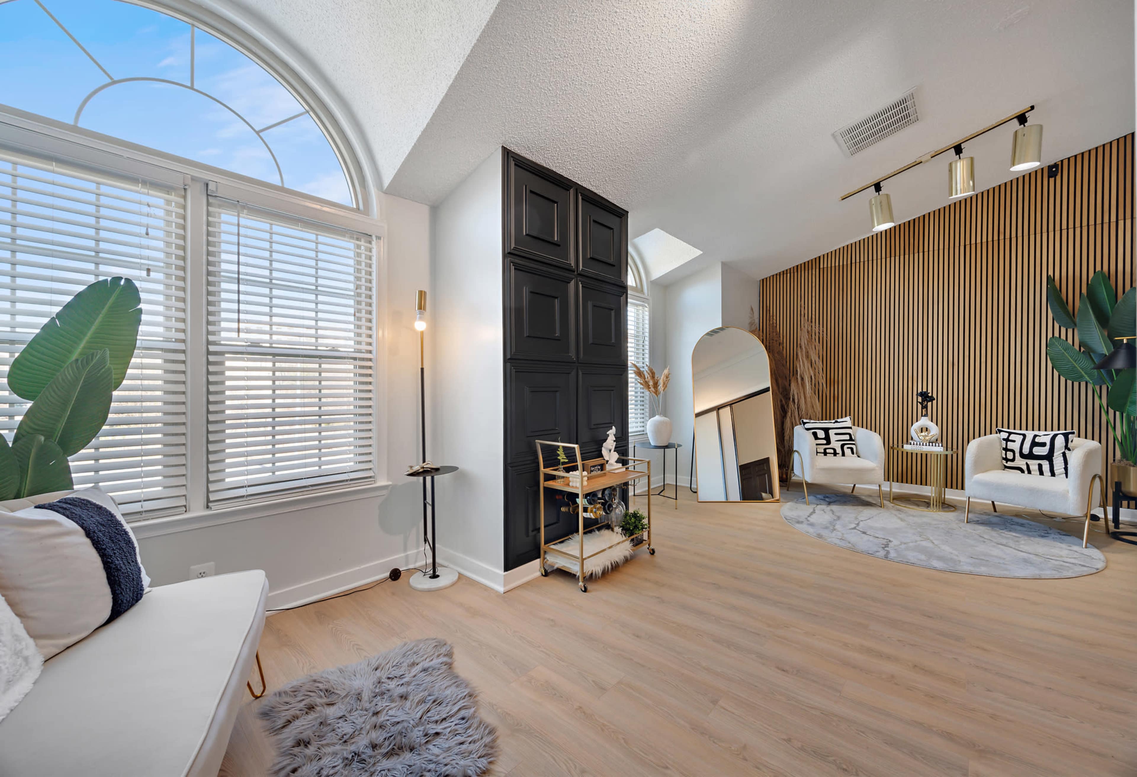 The scene features a bright room with large windows, light wood flooring, a circular mirror, and modern furnishings arranged against a backdrop of wooden paneling and plants.