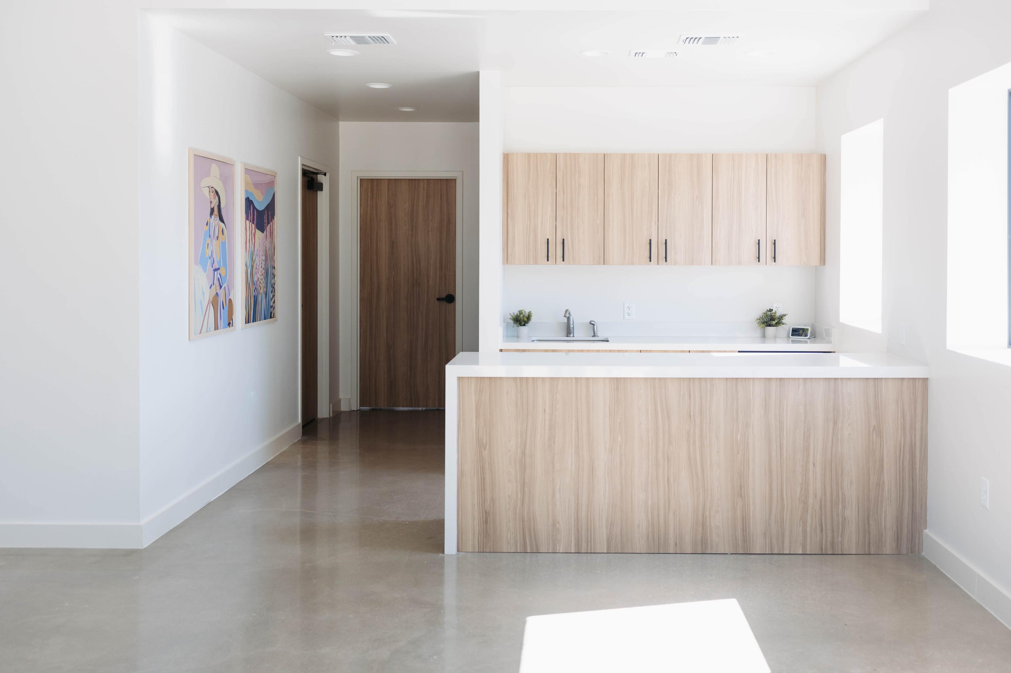 The image shows a modern, minimalist kitchen area with light wood cabinetry and white walls, adjacent to a hallway leading to a door.