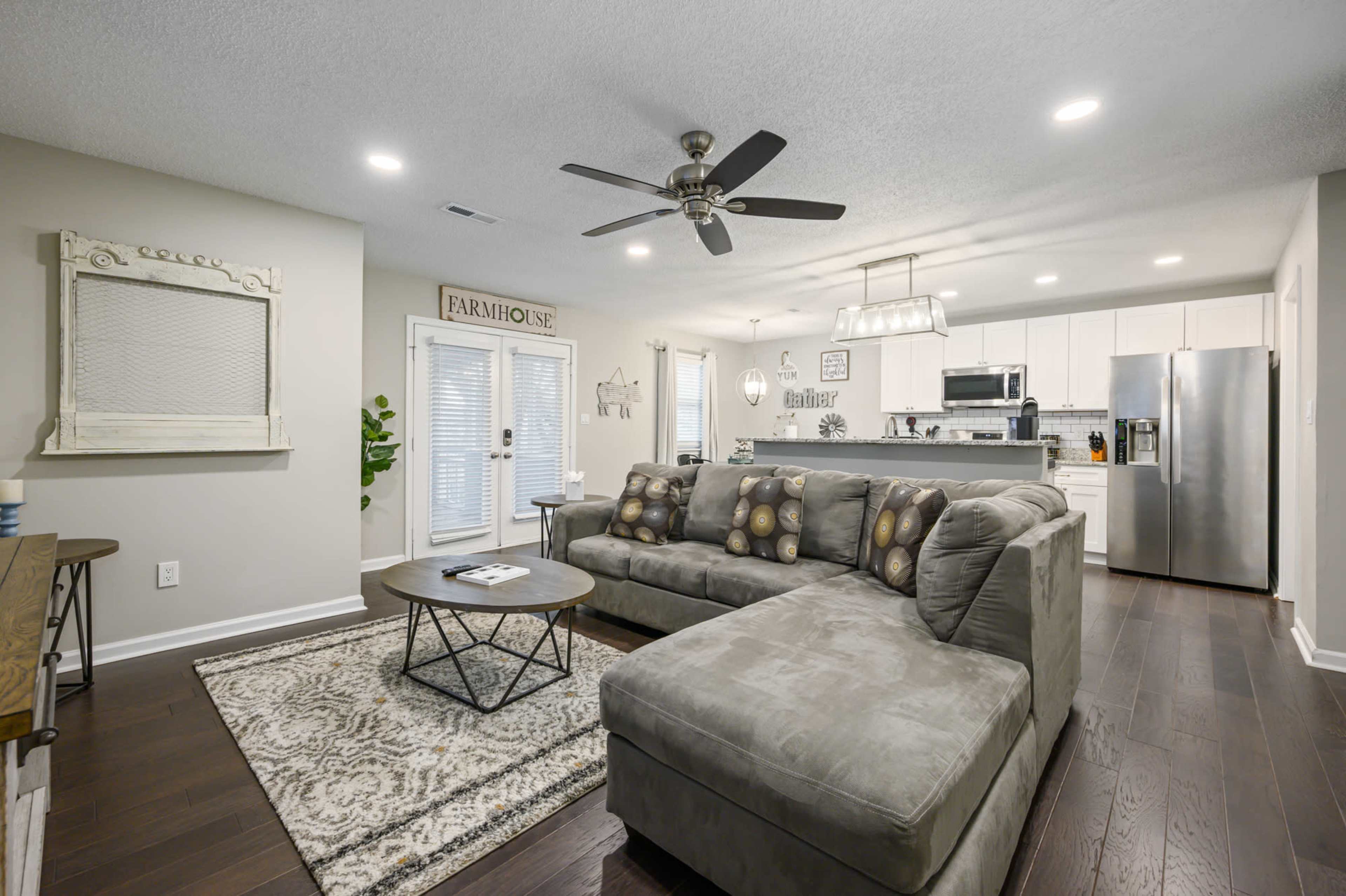 The image shows a modern living room featuring a gray sectional sofa, a round coffee table, and a kitchen area visible in the background.