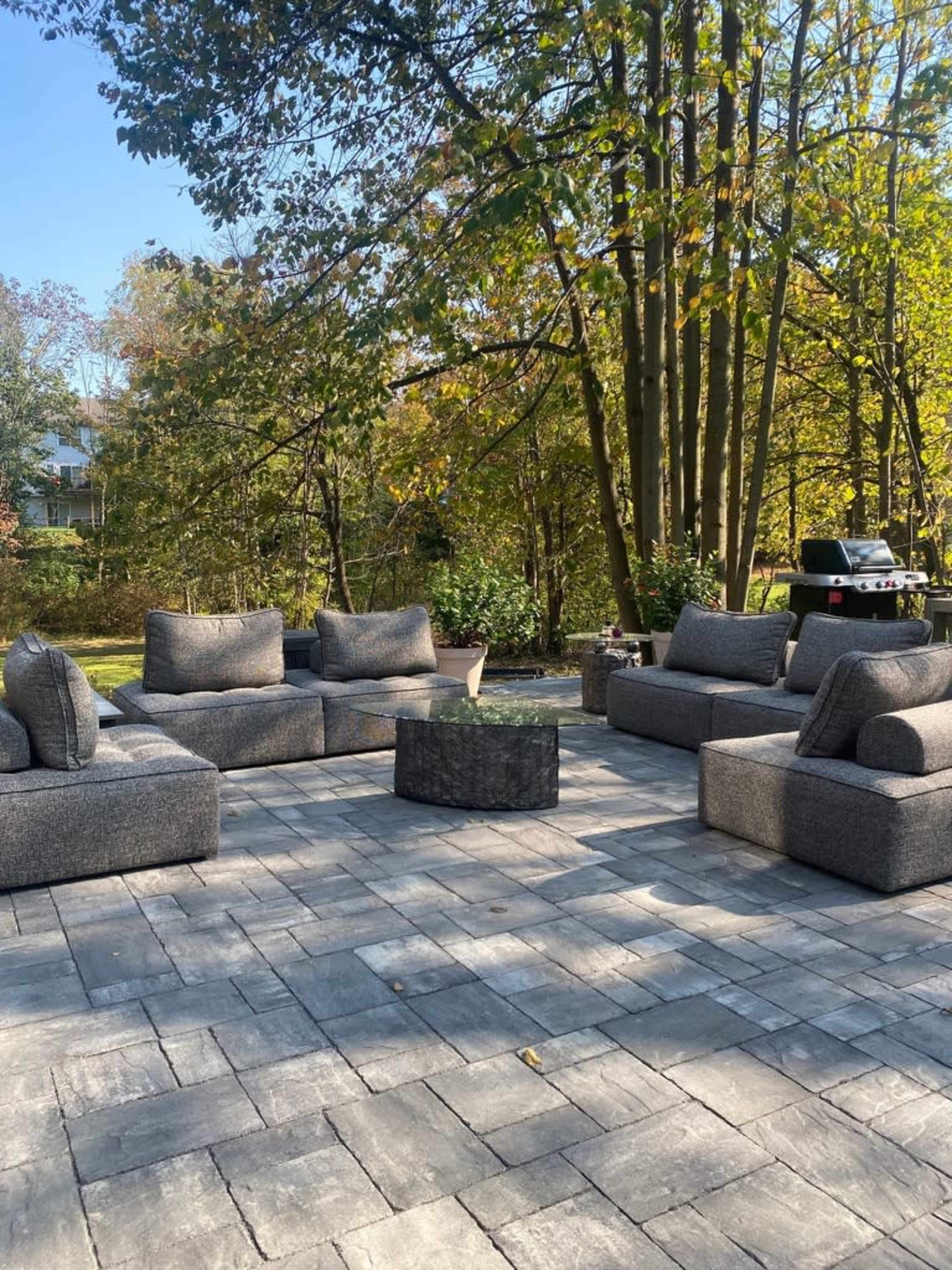 A patio seating area features several modular sofas arranged around a central table, surrounded by trees in a sunny outdoor setting.