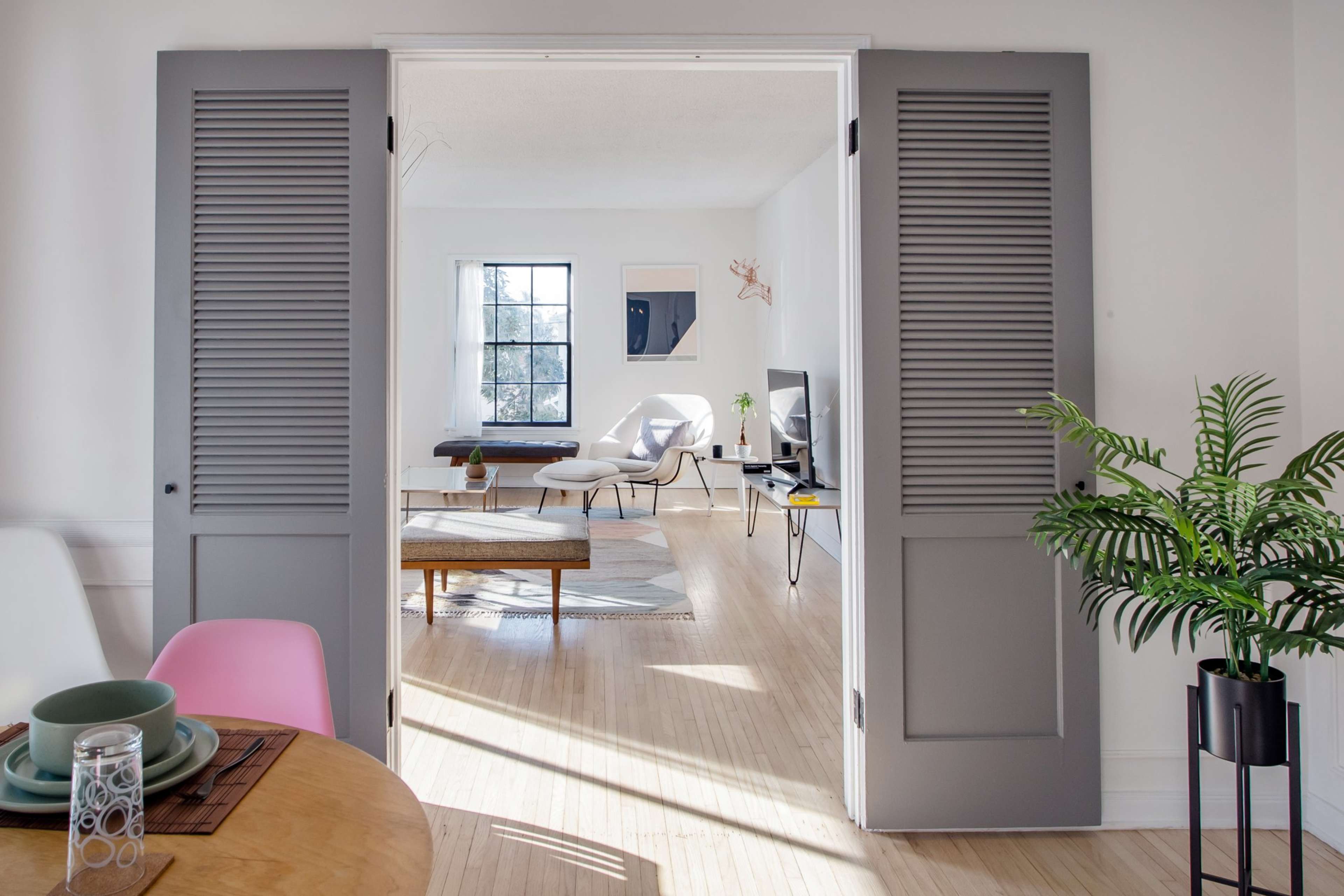 A doorway with shuttered doors leading into a bright living space featuring a minimalist design with a wooden dining table and a sitting area.