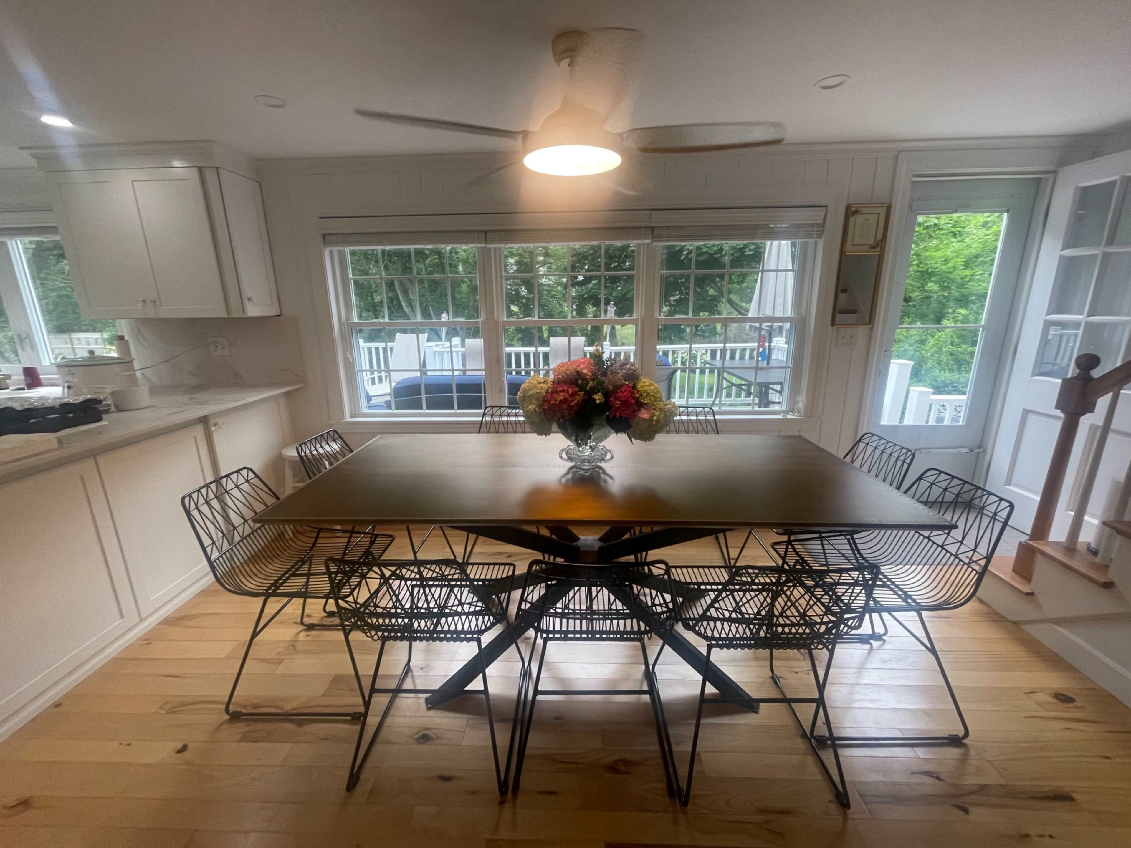 A dining area features a wooden table surrounded by wire-frame chairs, with a flower arrangement in the center and large windows overlooking greenery.