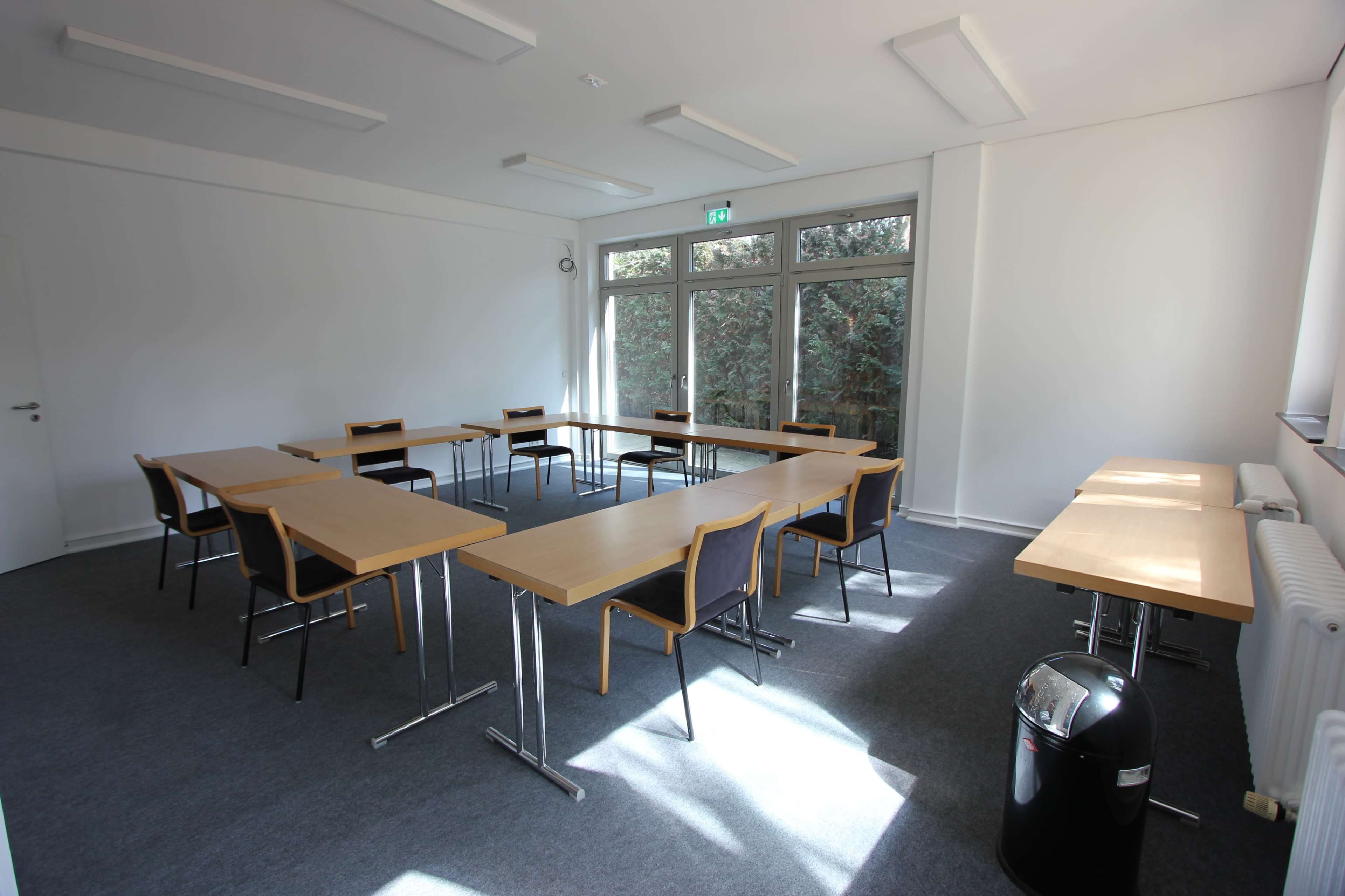 The image shows a spacious, well-lit classroom arranged with several wooden tables and chairs, and large windows providing a view of greenery outside.