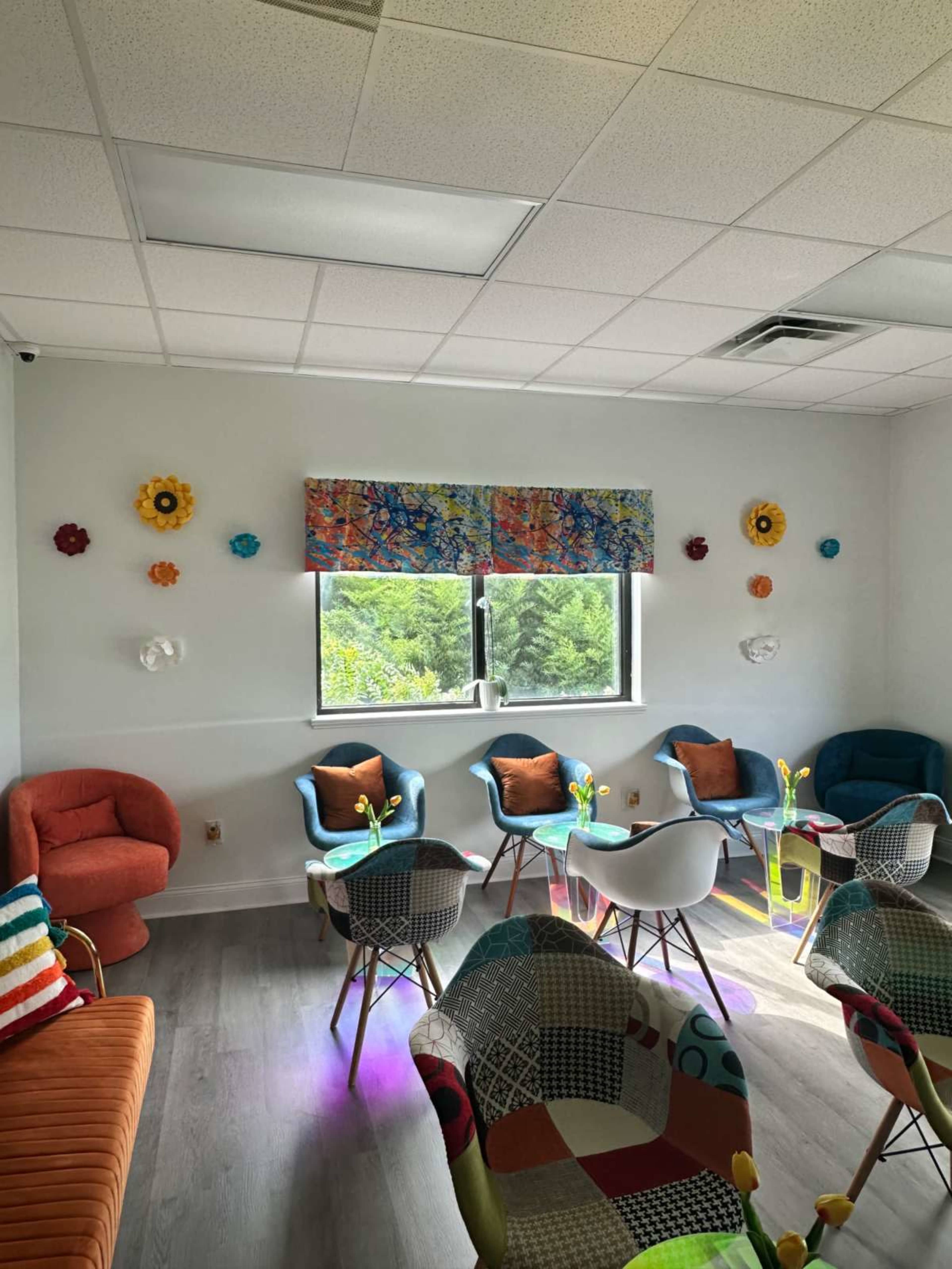 The image shows a brightly colored lounge area with various seating arrangements, floral wall decorations, and large windows letting in natural light.