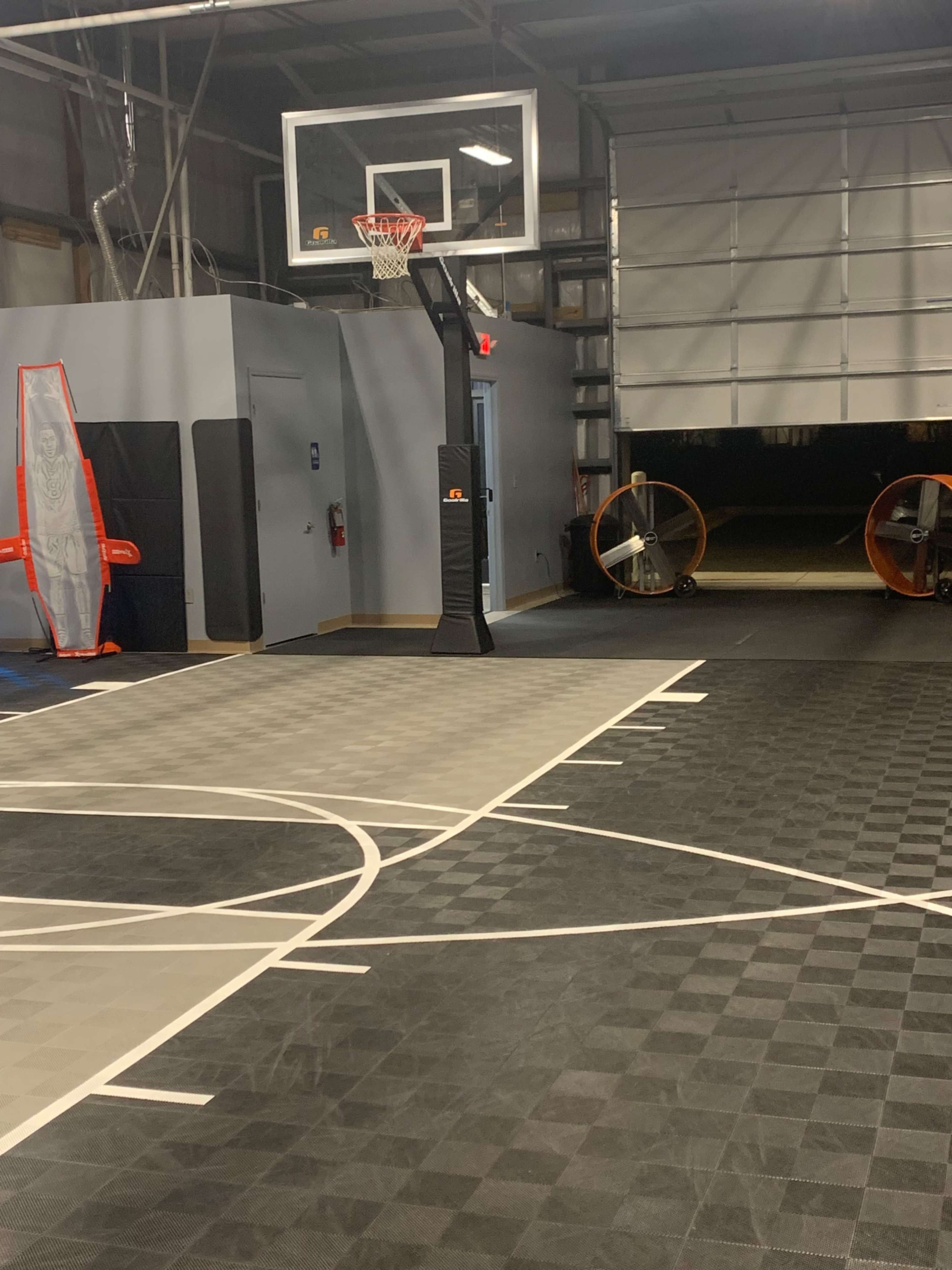 A basketball court with a hoop and a large overhead door opening to an outdoor area.