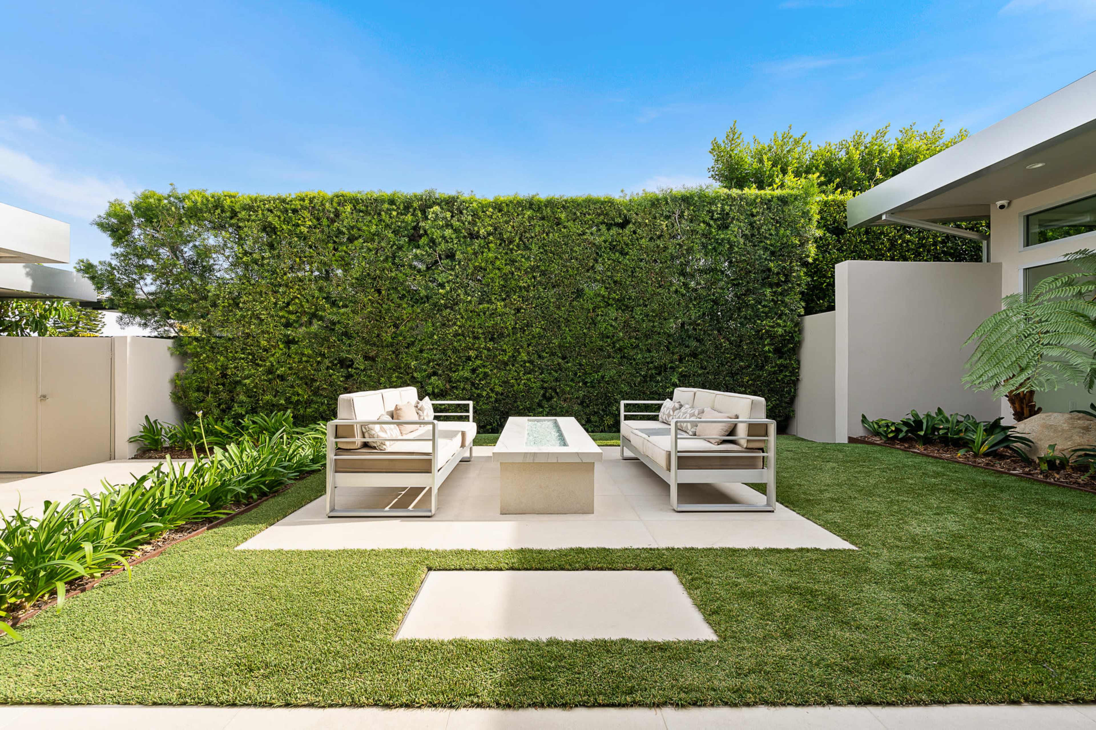 The image shows a neatly landscaped outdoor space featuring two modern sofas facing a rectangular coffee table on a grassy area, surrounded by lush greenery and a minimalist architectural backdrop.