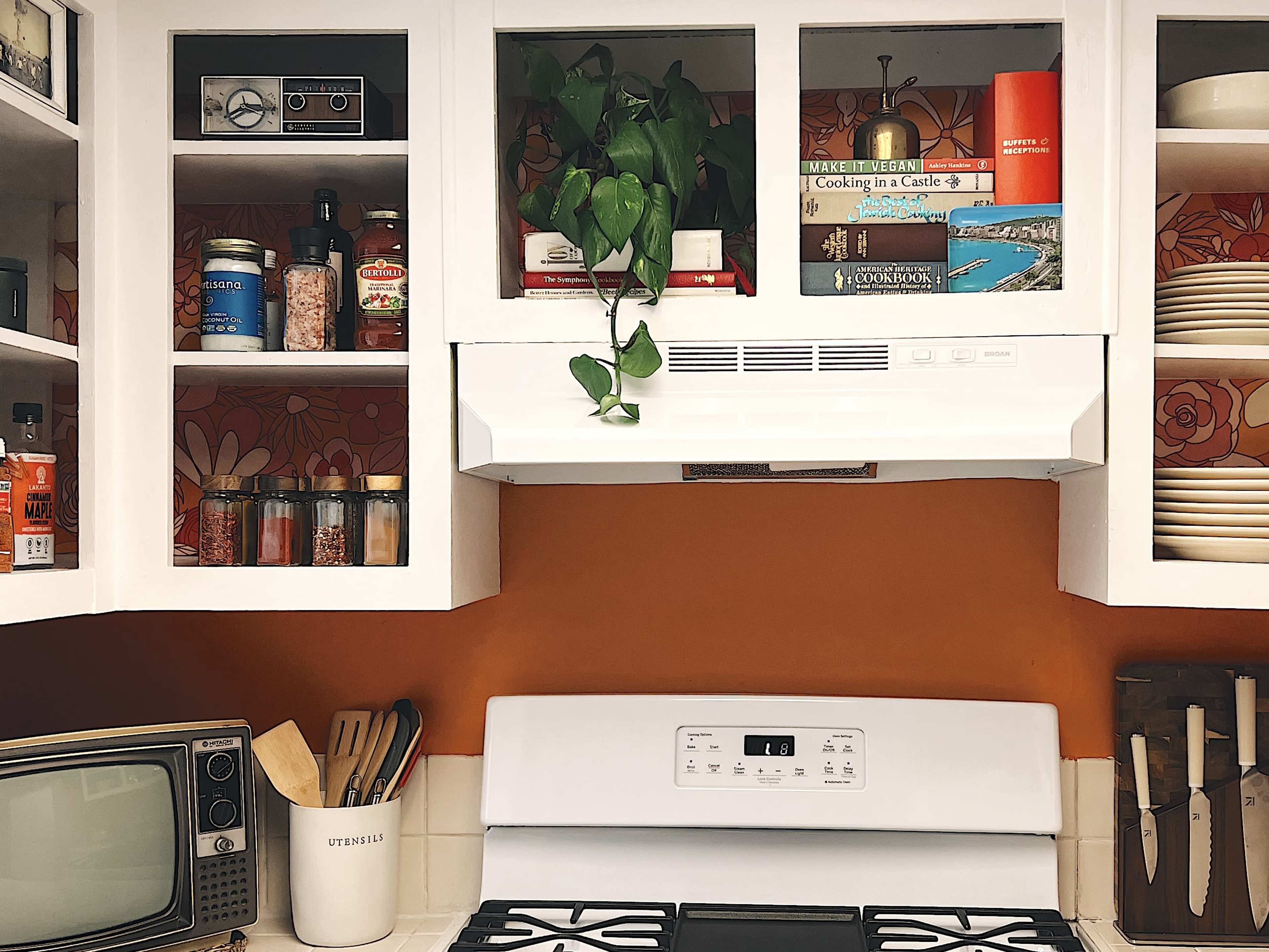 A kitchen scene features white cabinets with glass doors displaying jars and books, a white stove beneath a hood, and a vintage television on the counter.