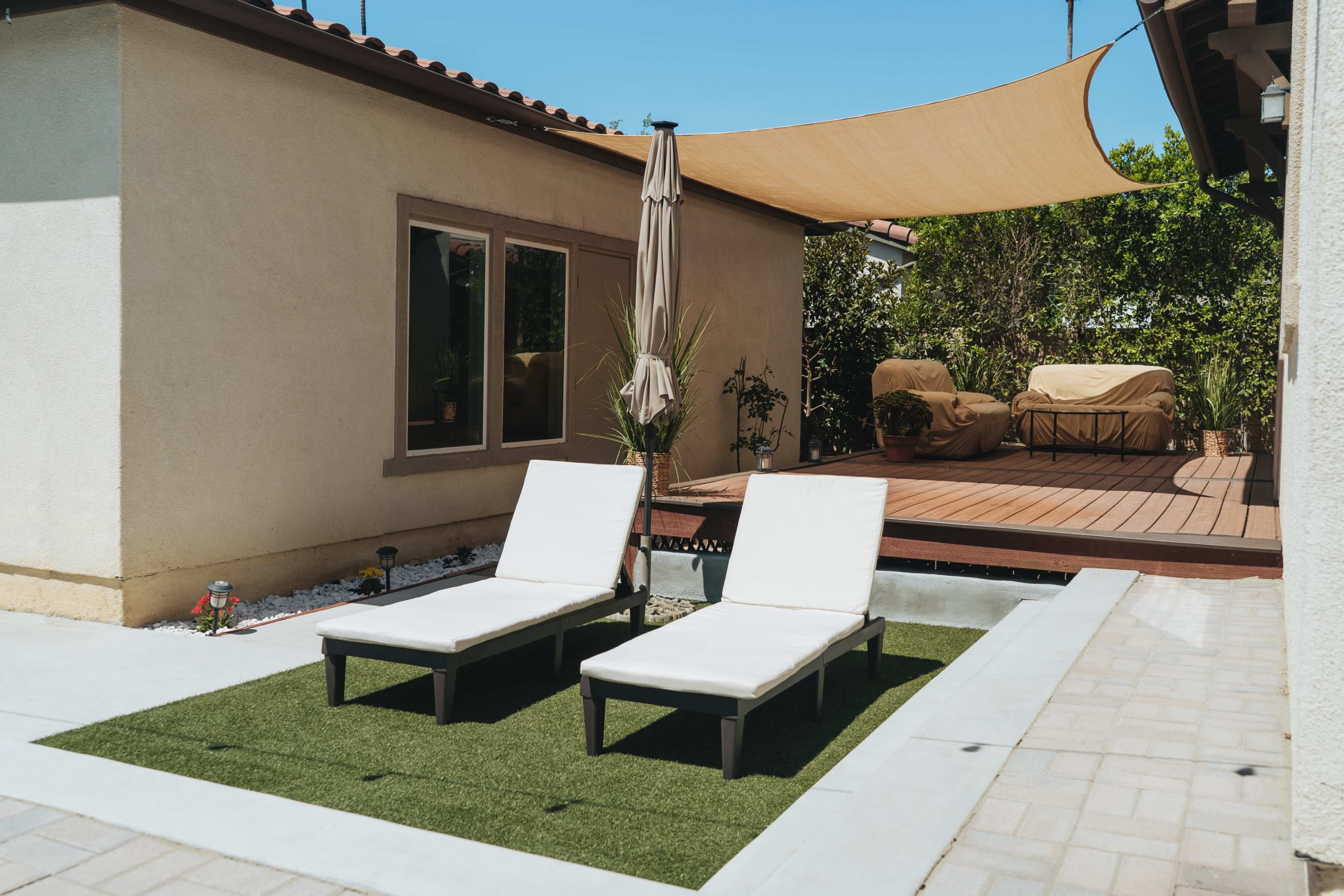 Two white lounge chairs on a patch of artificial grass in a patio area with a sunshade and a wooden deck in the background.