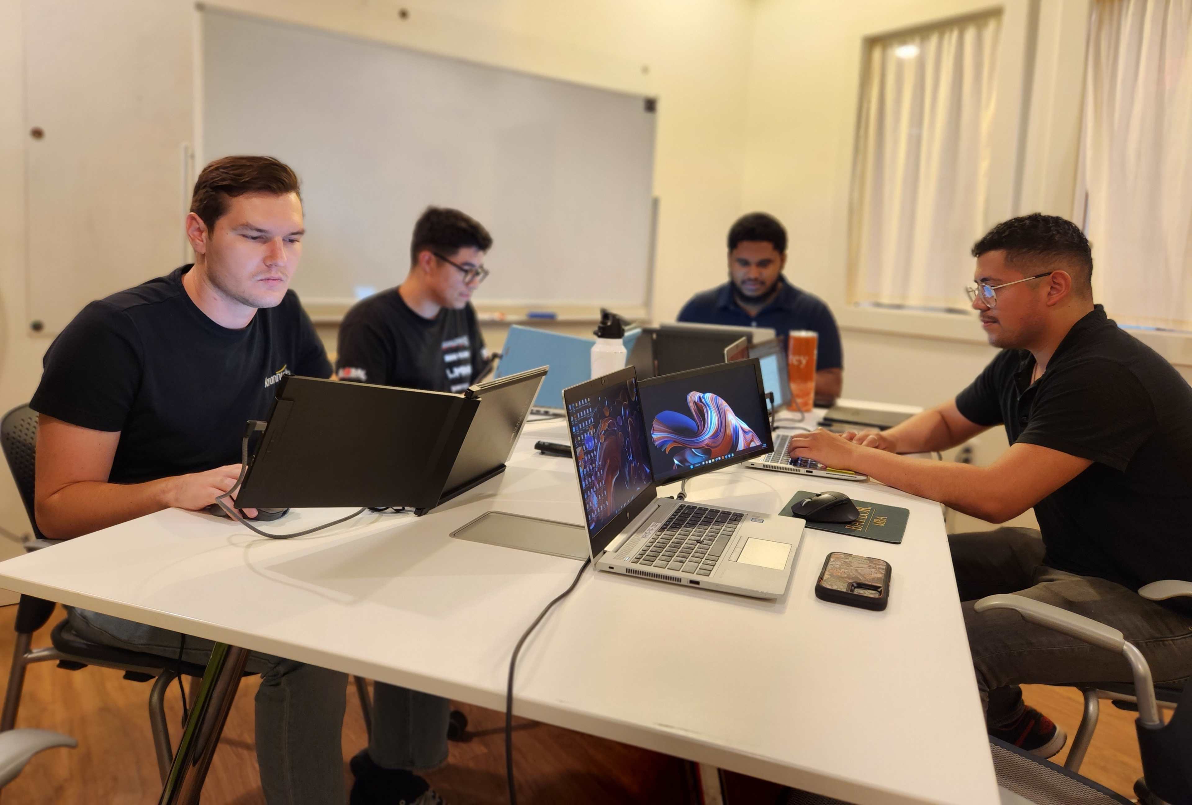 Four individuals are seated around a table, each working on their laptops in a well-lit room with a whiteboard in the background.