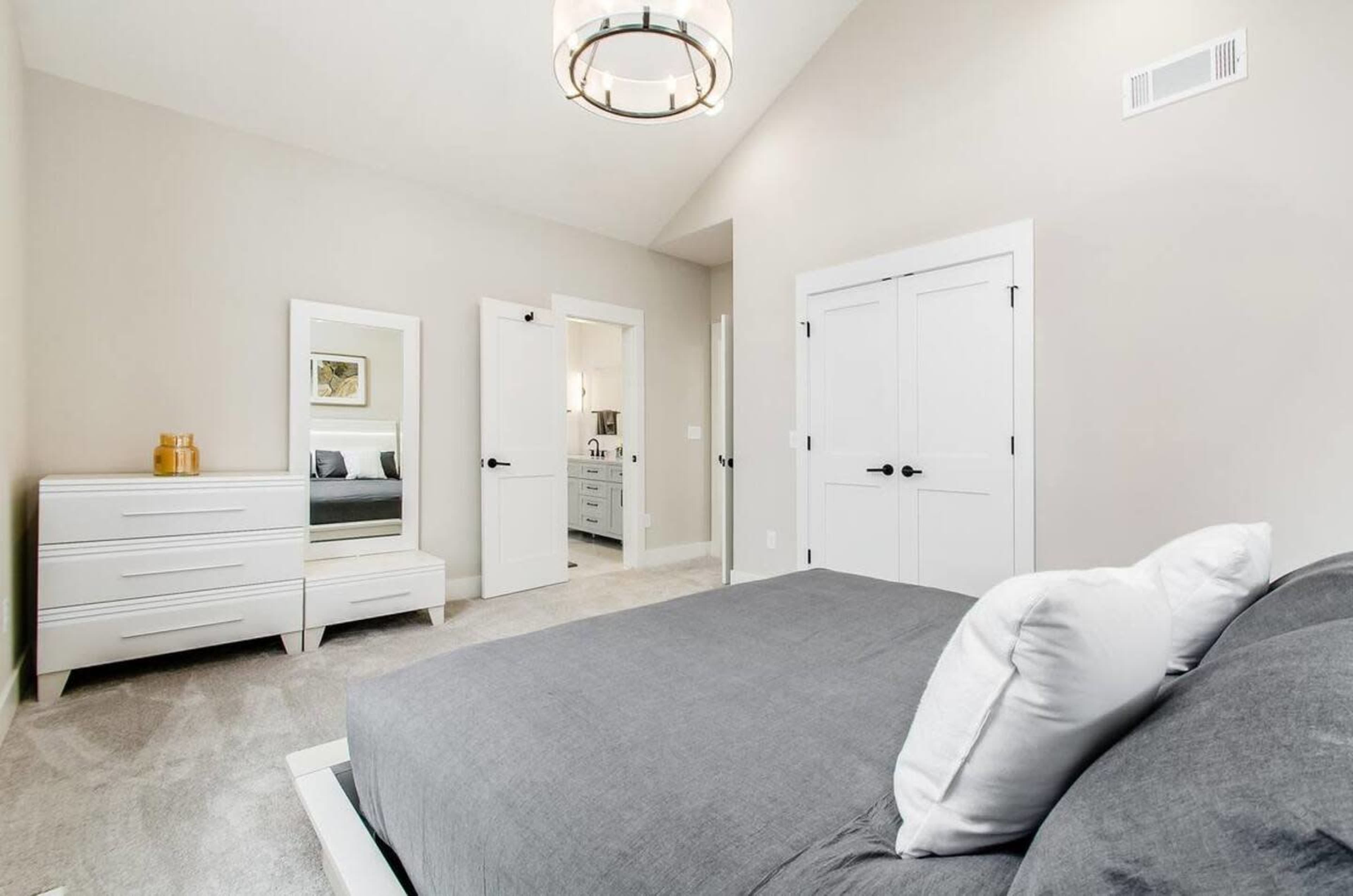 The image shows a modern bedroom with a gray bedspread, a dresser, a mirror, and two white doors leading to another room.