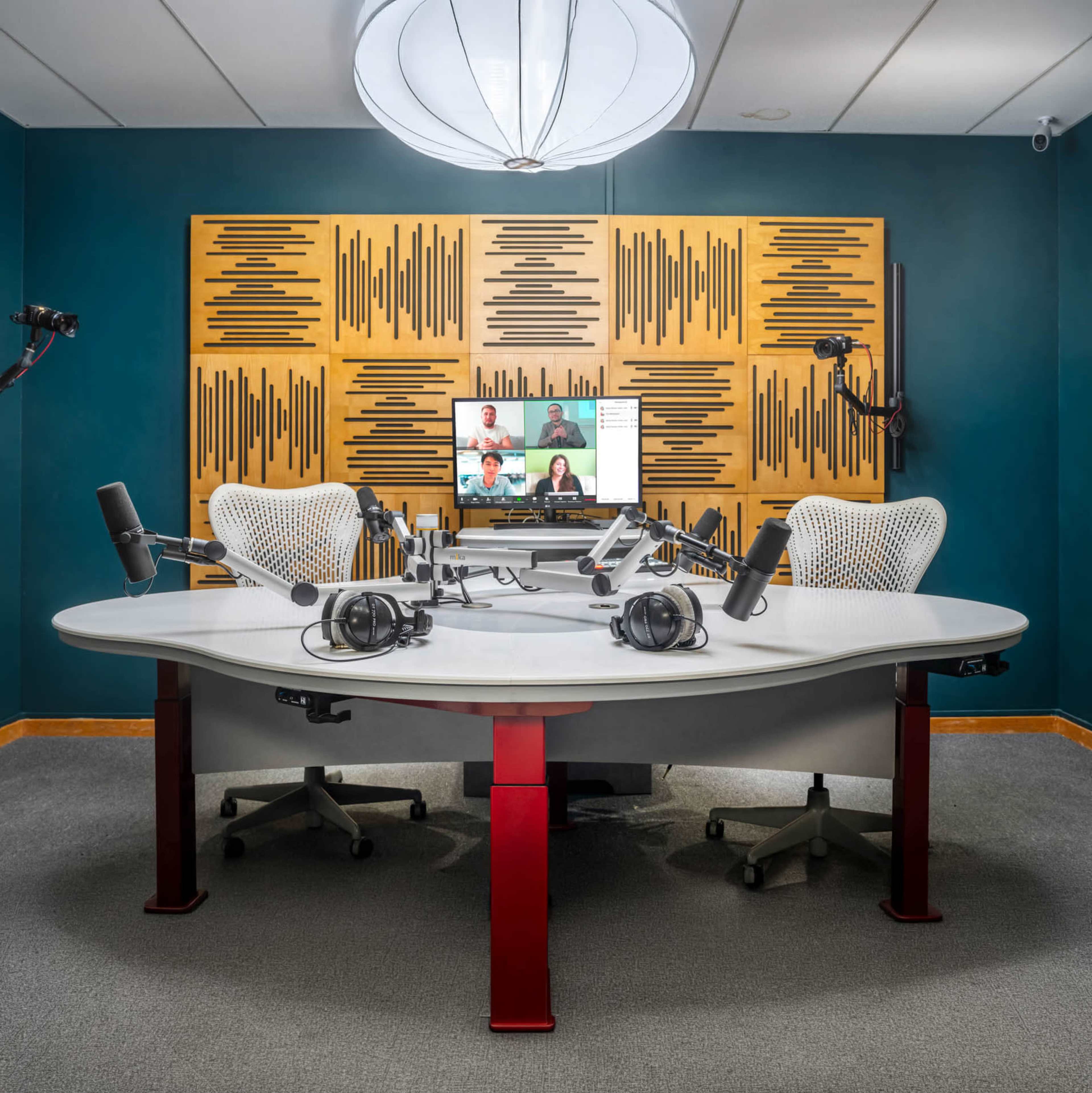 The image shows a modern podcast studio with a large oval table, multiple microphones, headphones, and a screen displaying participants in a virtual meeting.