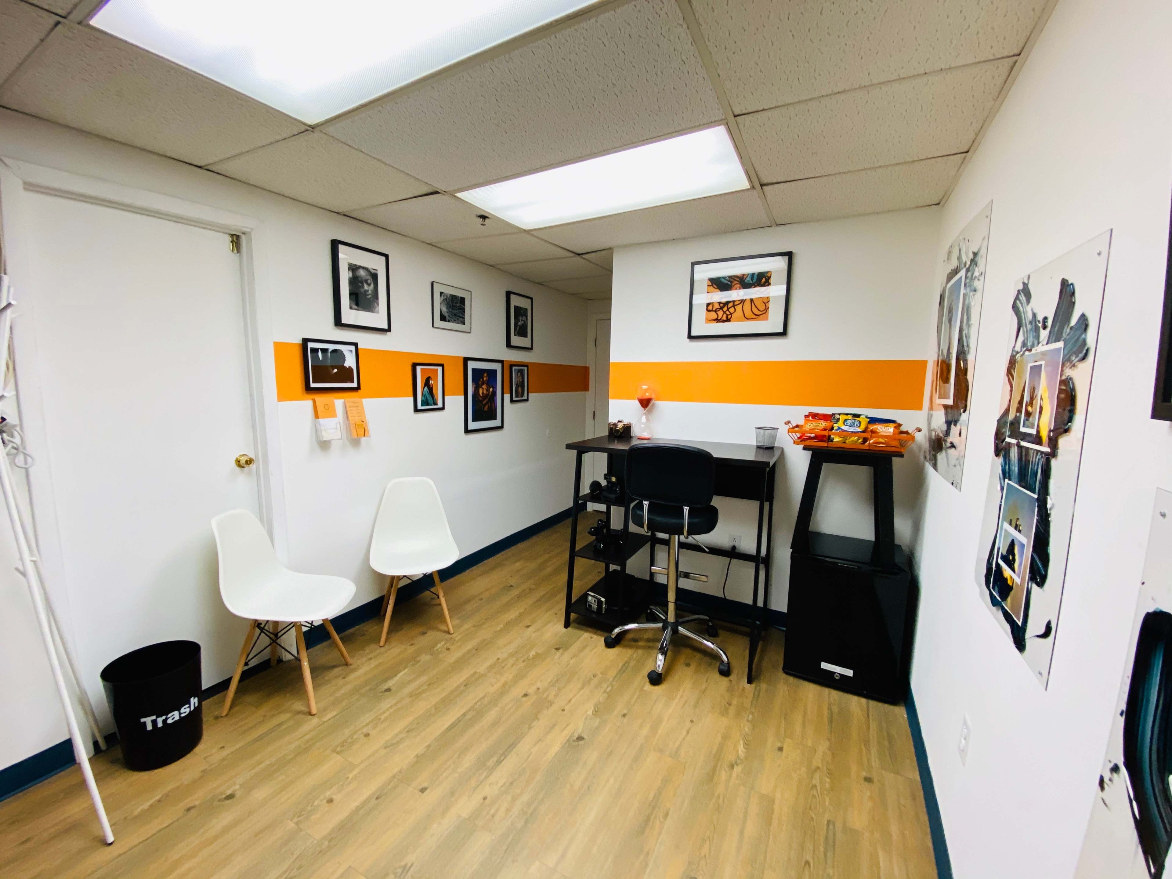 A small office space with two white chairs, a black desk and chair, and framed photos on the walls, featuring an orange accent stripe.