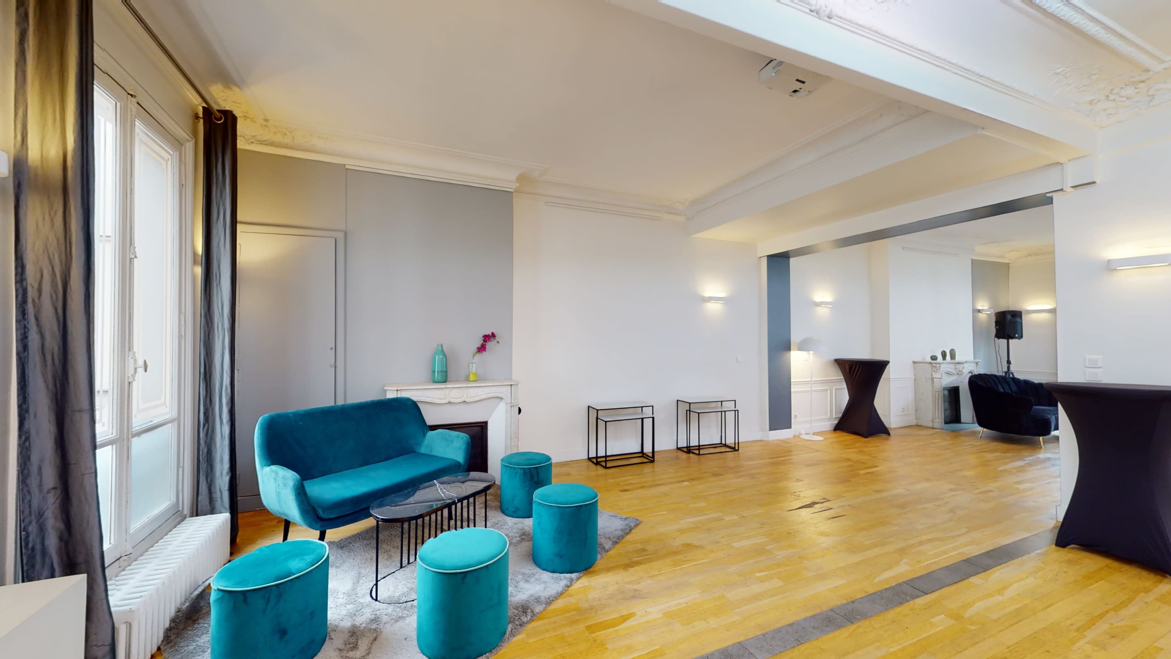 A modern interior space with a teal sofa, circular tables, and sleek chairs, featuring large windows and hardwood flooring.