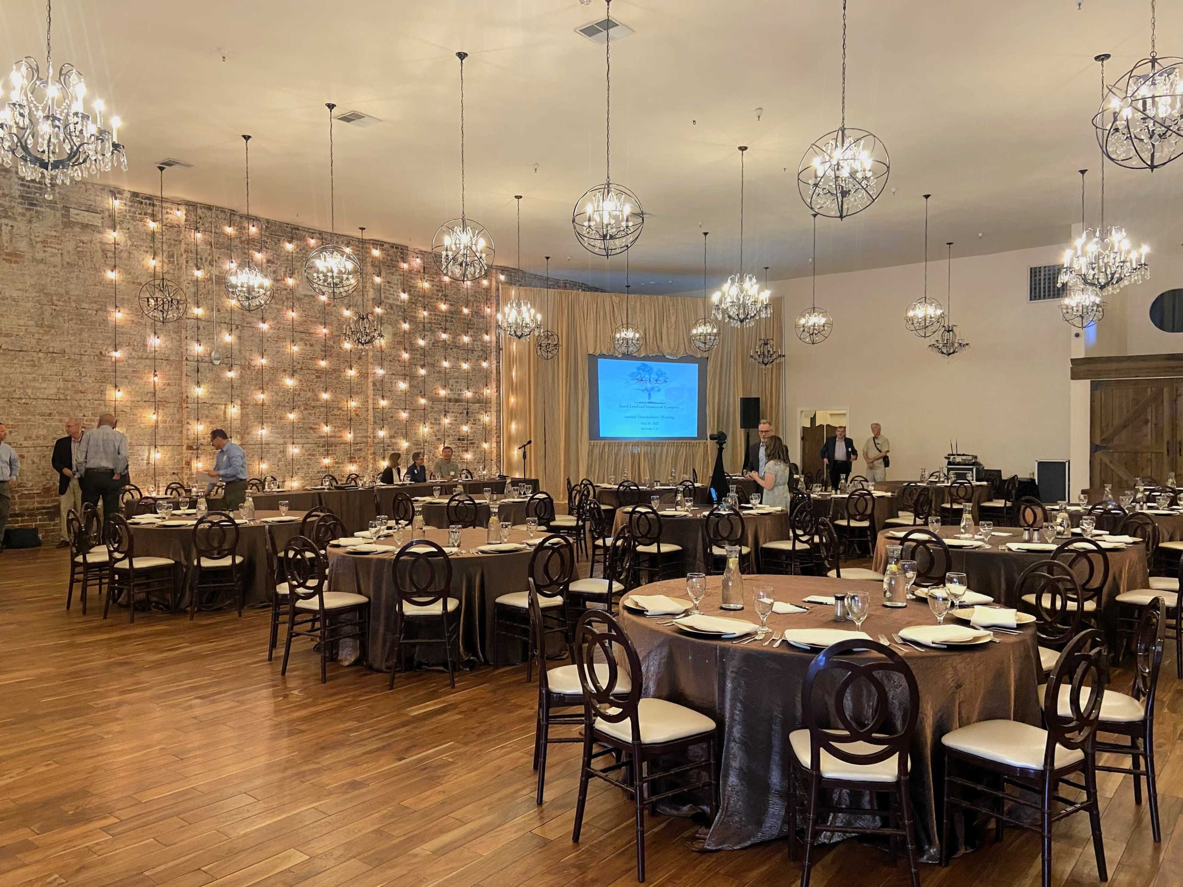 A spacious banquet hall set up with round tables and illuminated chandeliers, featuring a backdrop of string lights.