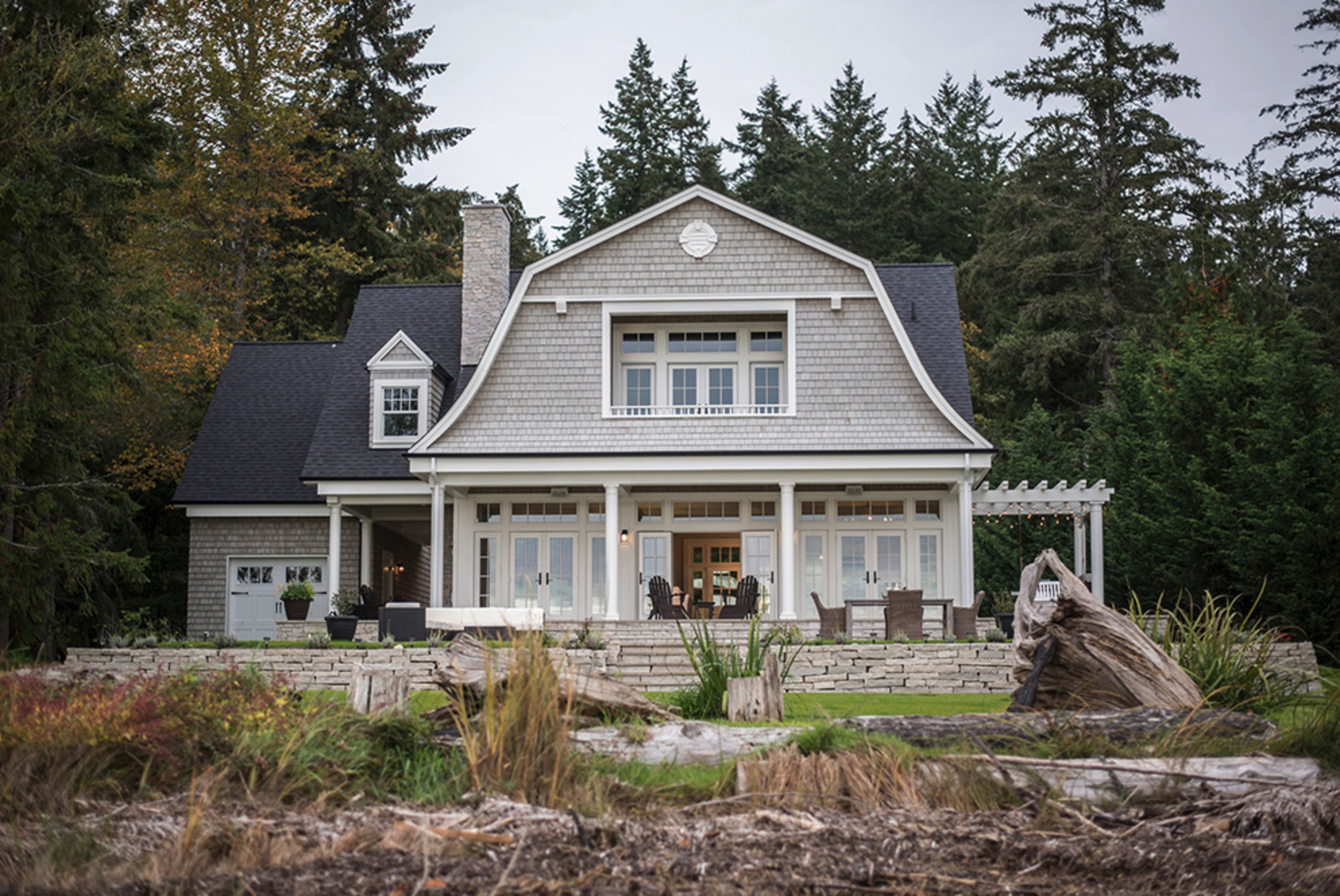 A coastal-style house with a gray shingle exterior features a spacious front porch and is surrounded by trees and landscaping.