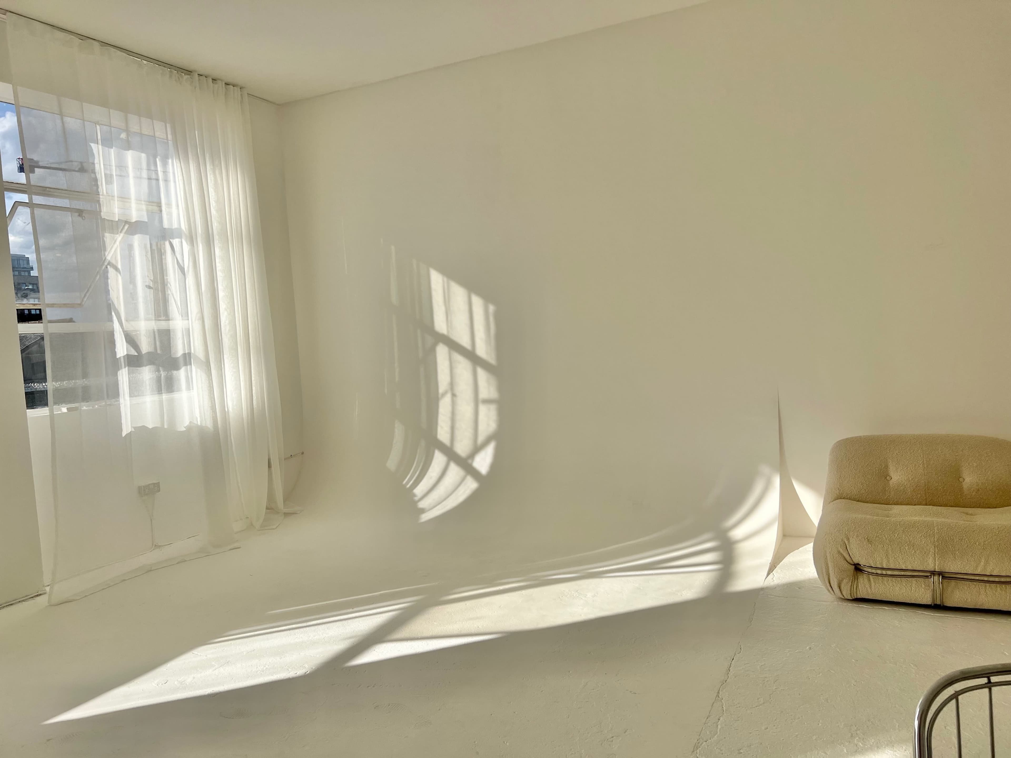 A spacious, bright, white room with a large window allowing sunlight to create shadows on the floor, and a beige couch positioned against one wall.