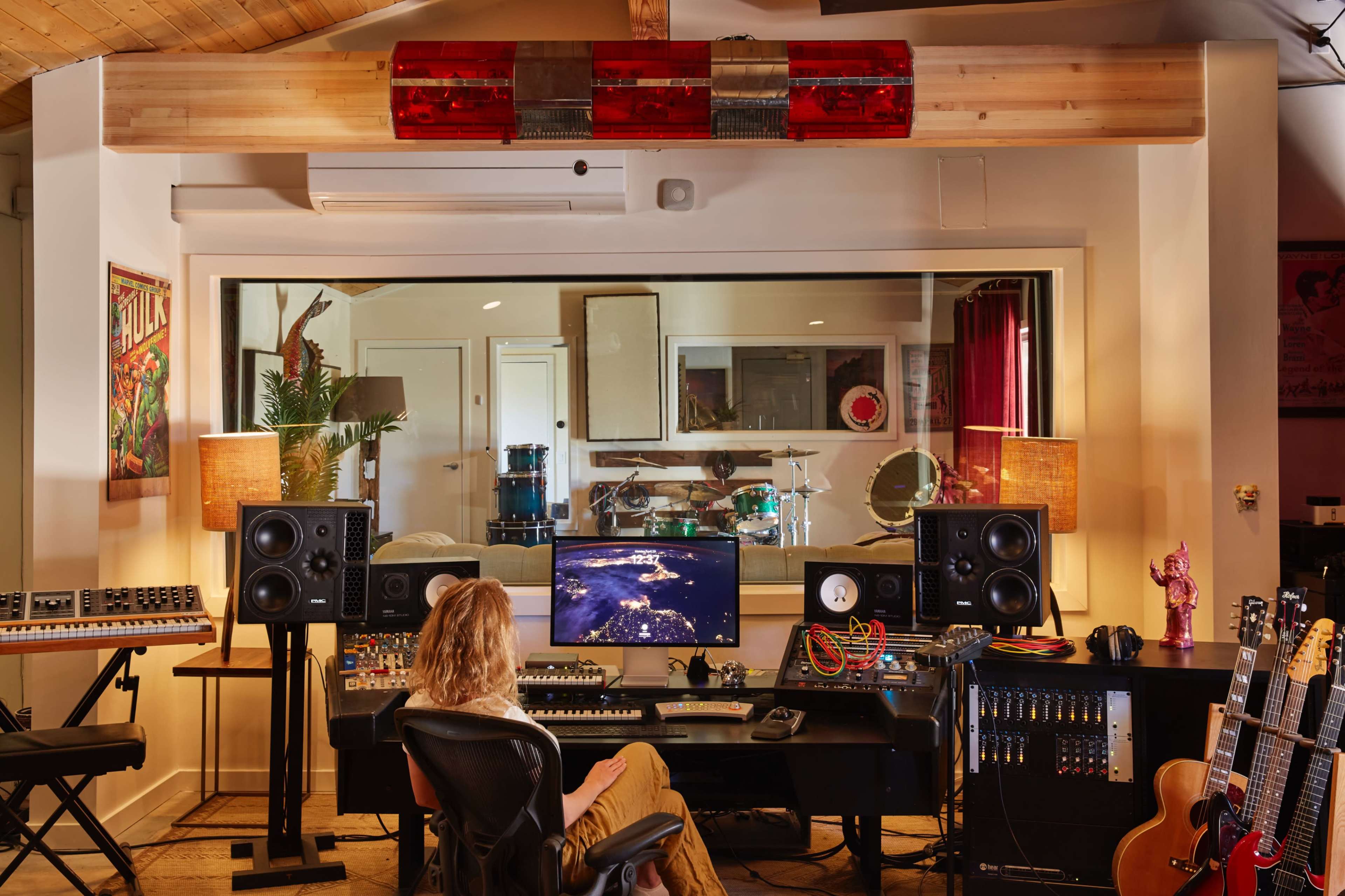 A person sits in a chair at a music production desk surrounded by audio equipment and monitors in a well-lit recording studio.