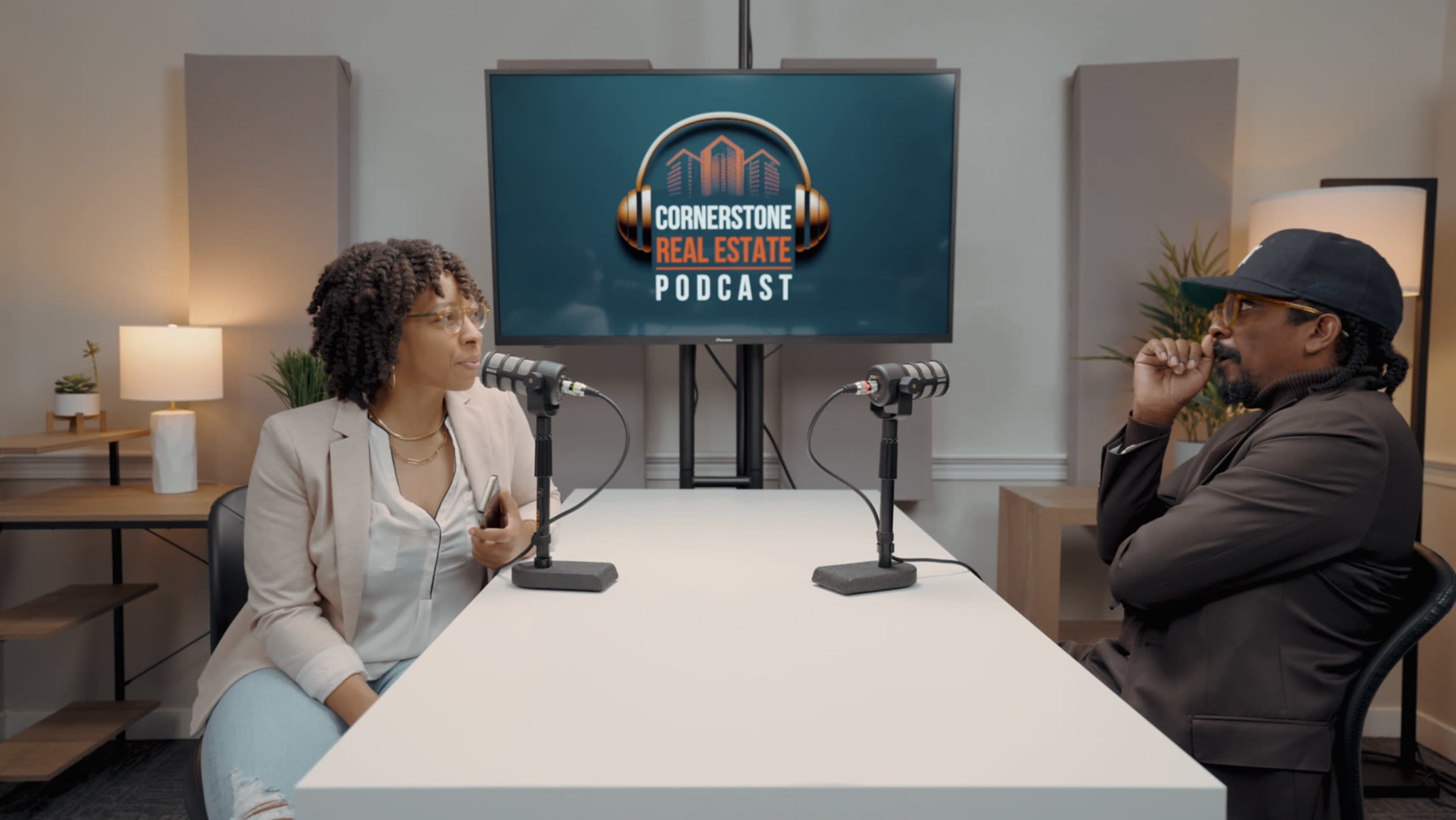 A woman and a man sit across from each other at a table equipped with microphones in a podcast studio, with a backdrop displaying the "Cornerstone Real Estate Podcast" logo.