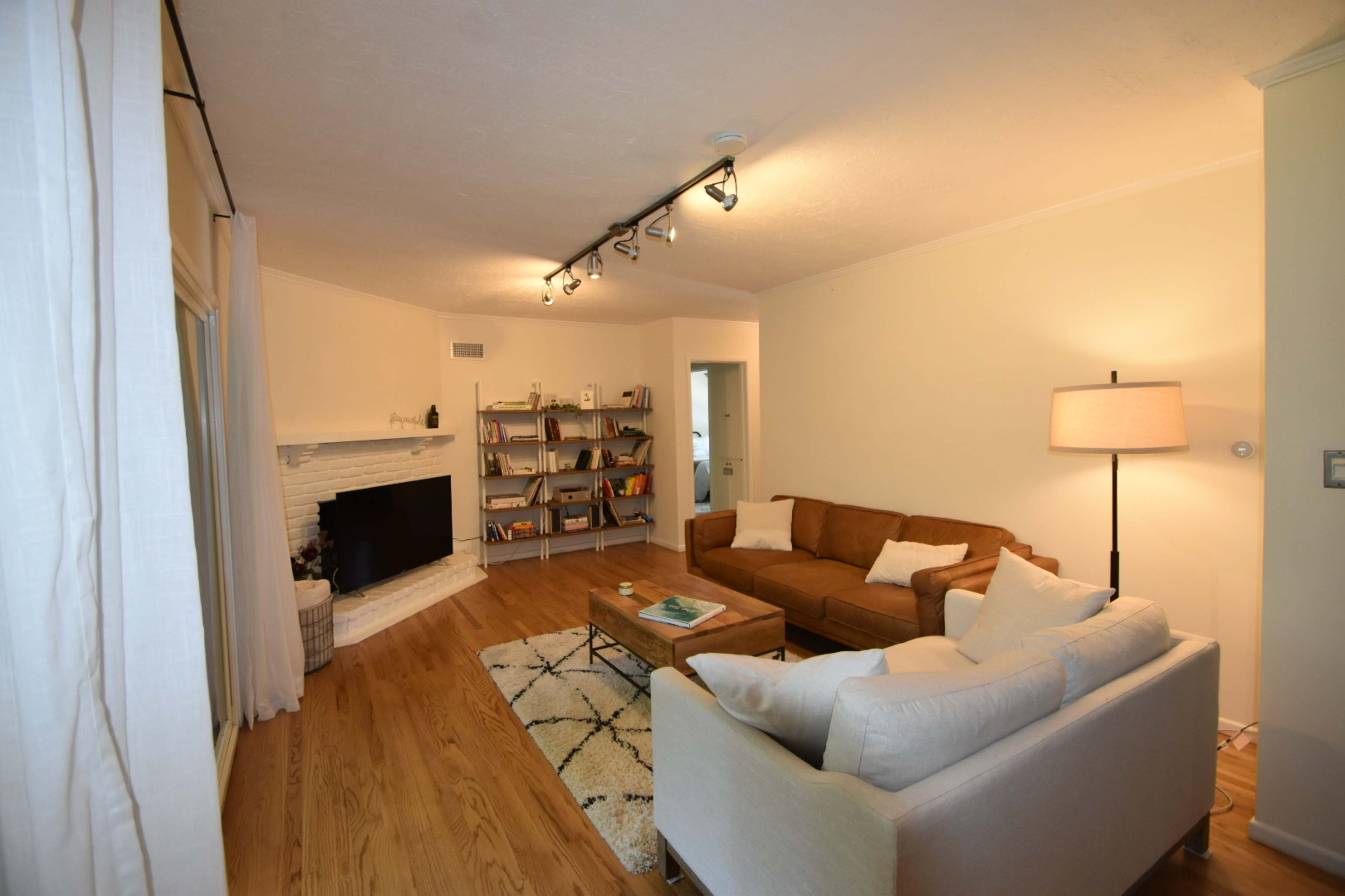 The living room features a brown sectional sofa, a white couch, a wooden coffee table, a bookshelf filled with books, and a fireplace with a white brick surround.