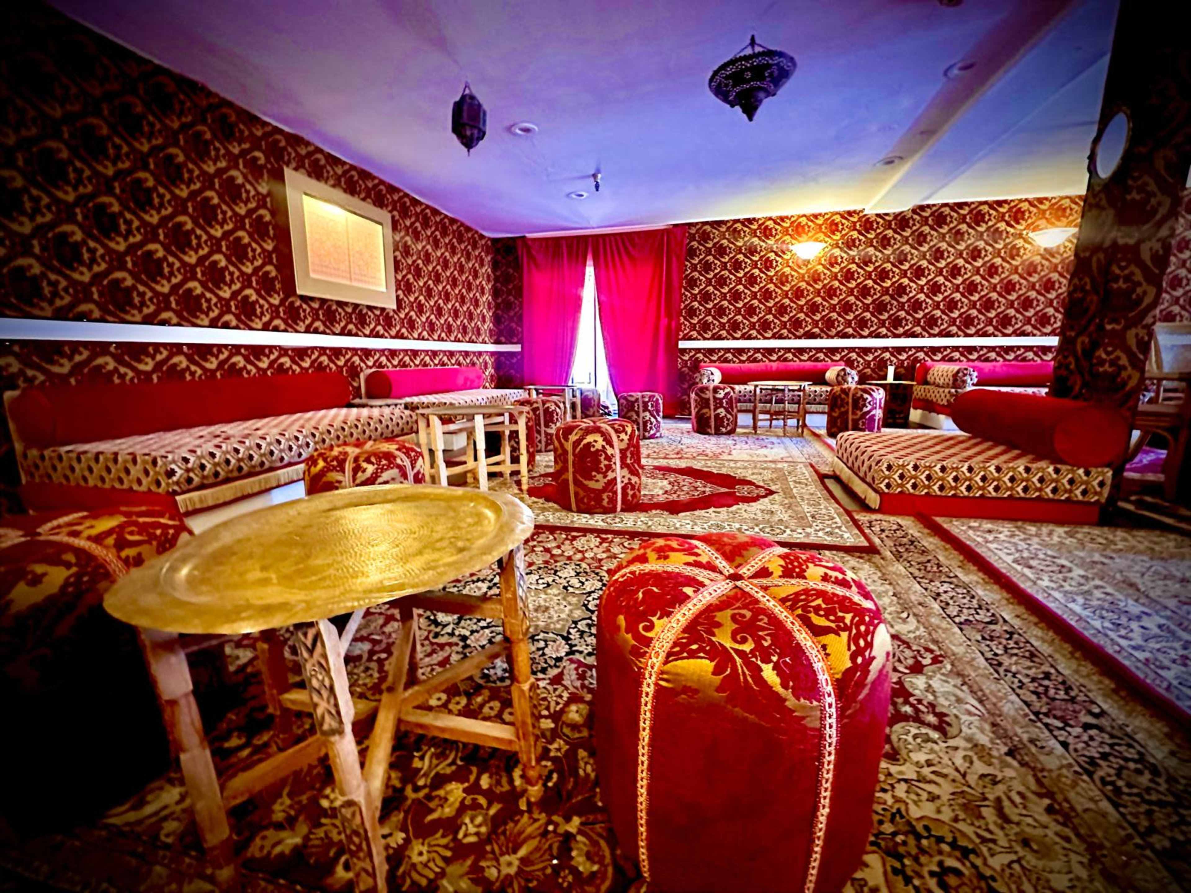 The image shows a well-decorated room featuring ornate red-patterned furnishings, with low seating and intricate rugs covering the floor.