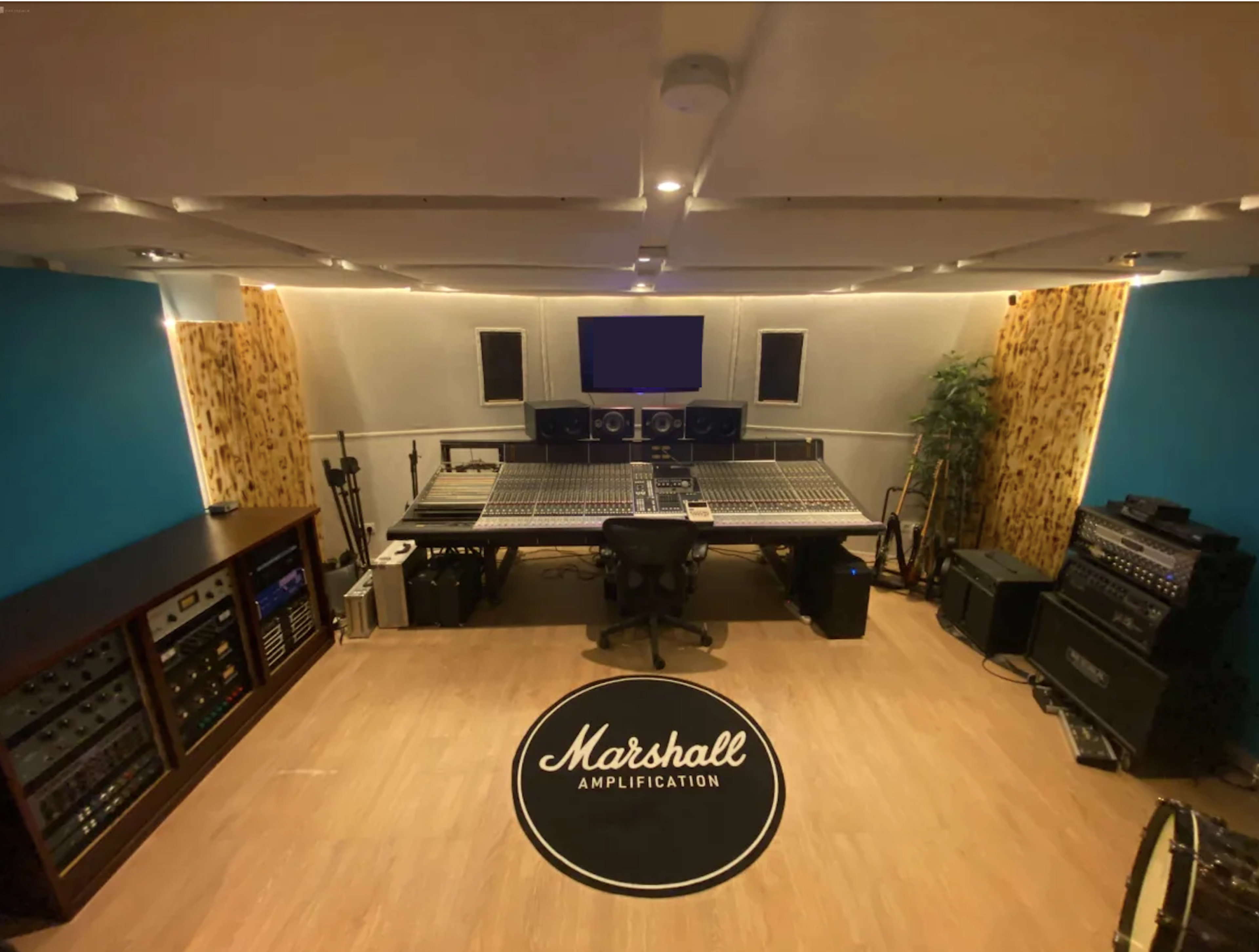 A music recording studio with a central mixing console, surrounded by audio equipment and a large logo on the floor.