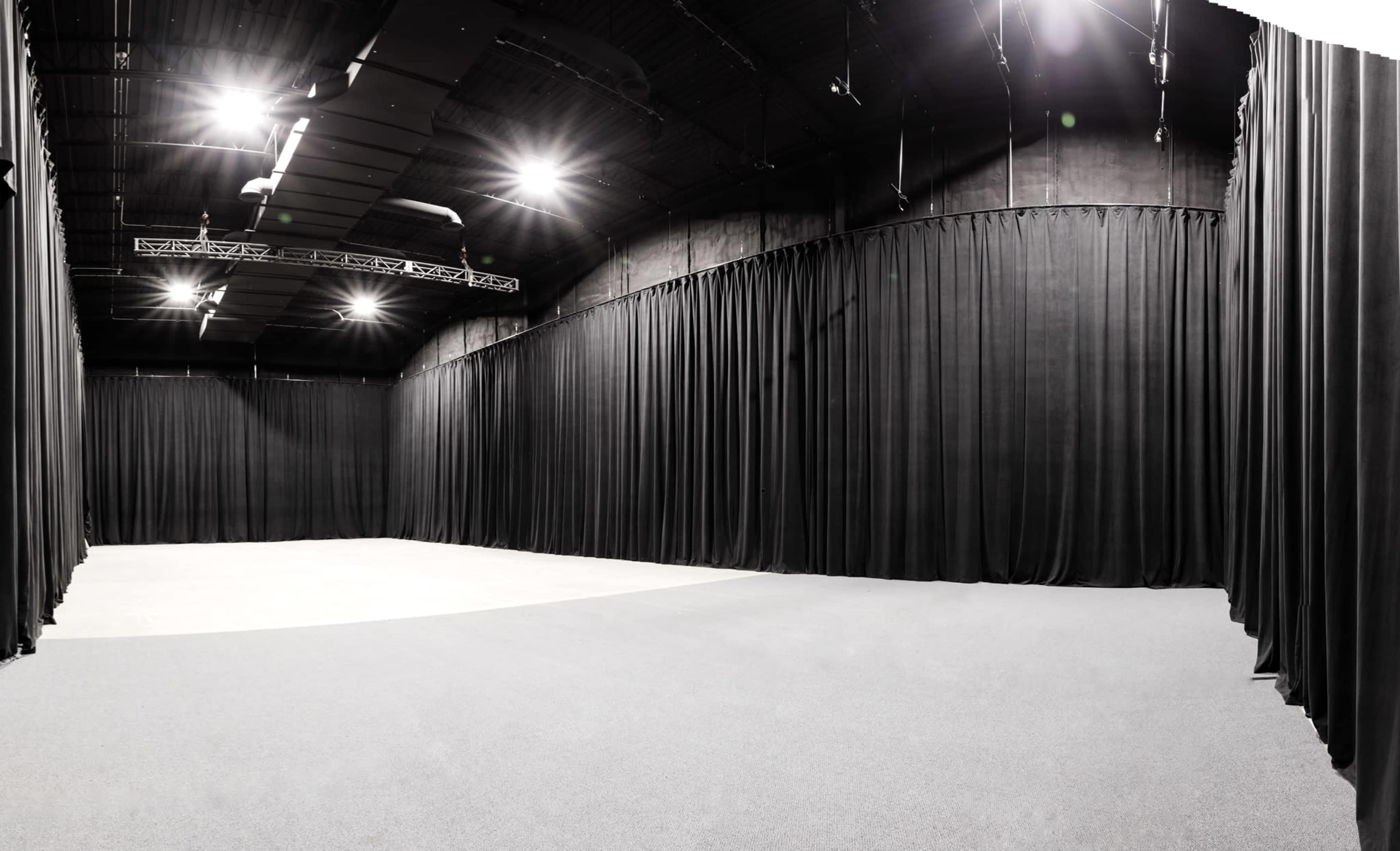 A spacious, empty studio with black curtains lining the walls and bright overhead lights illuminating a gray carpeted floor.