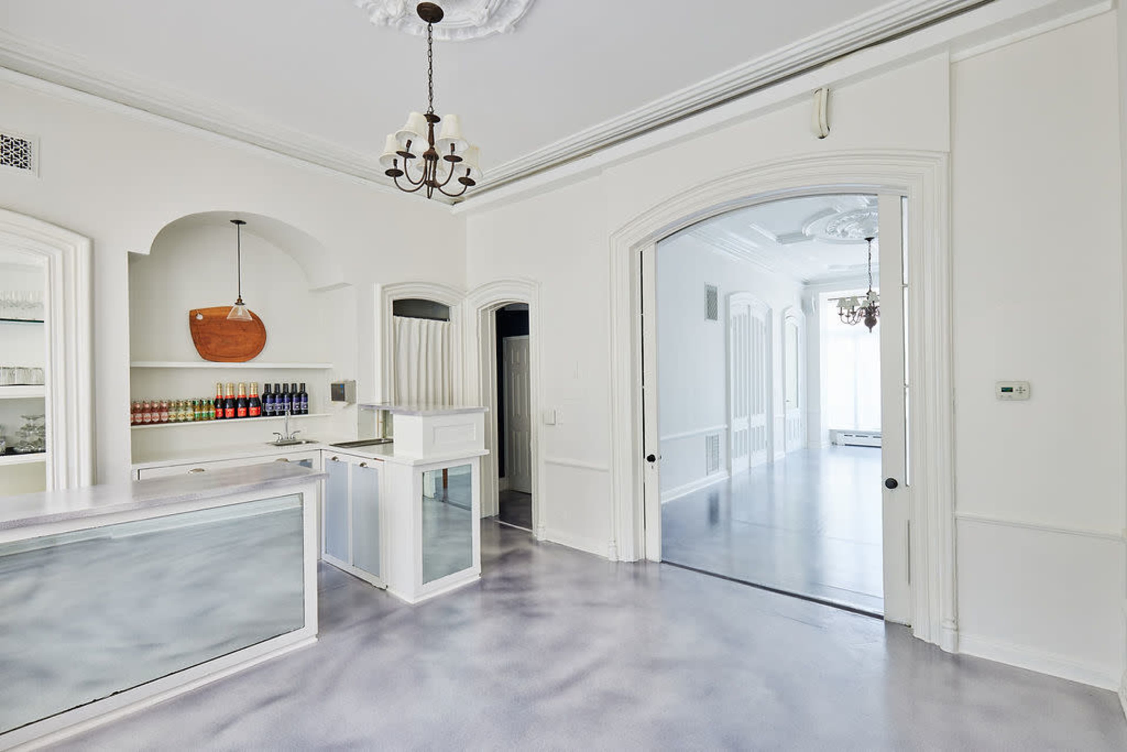 A bright, minimalist interior features a reception area with a counter, shelves stocked with beverages, and large archways leading to adjoining rooms.