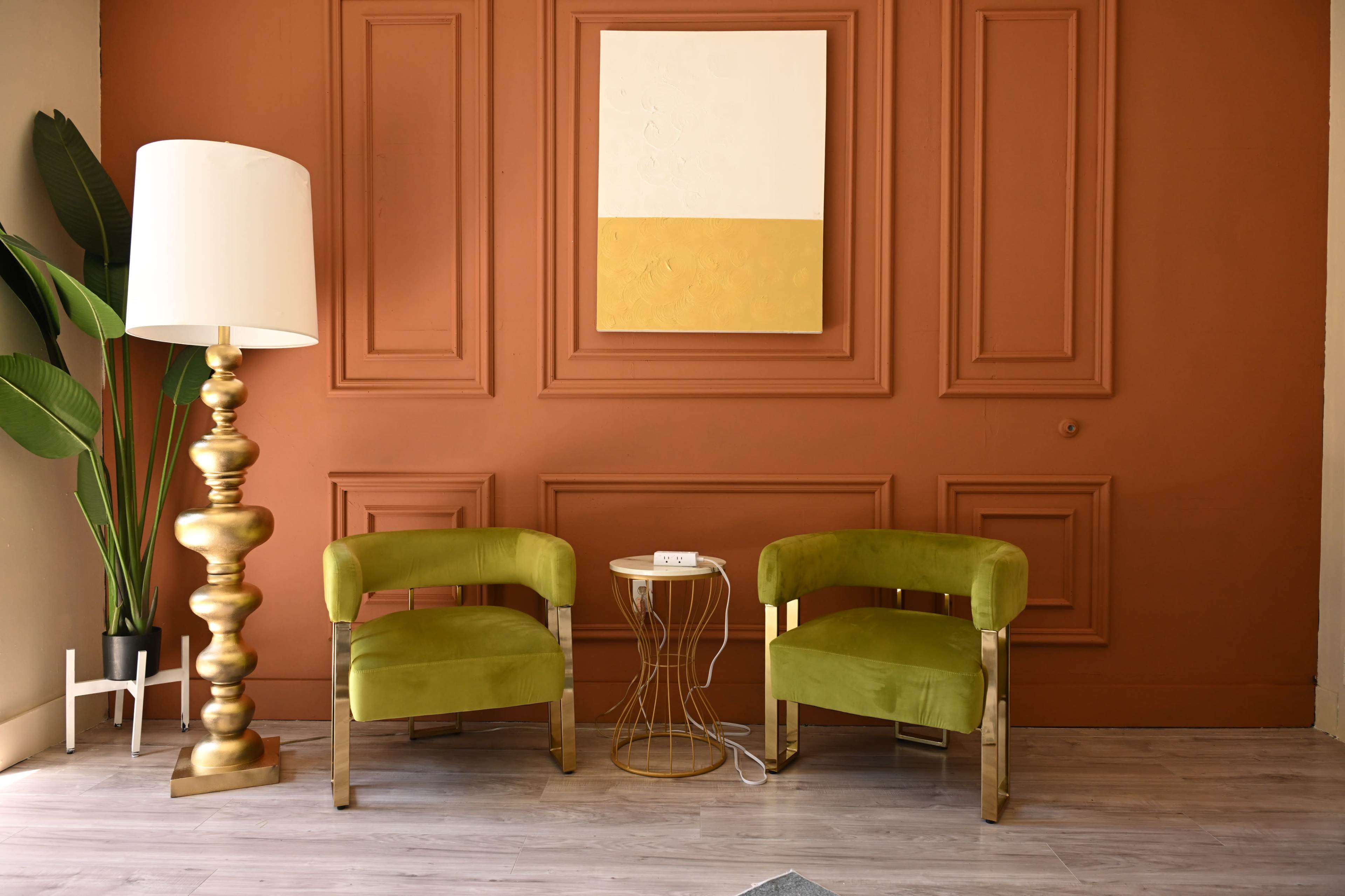 The image features a cozy sitting area with two green chairs, a decorative gold lamp, and a small side table against a rust-colored wall with a large, simple artwork above.