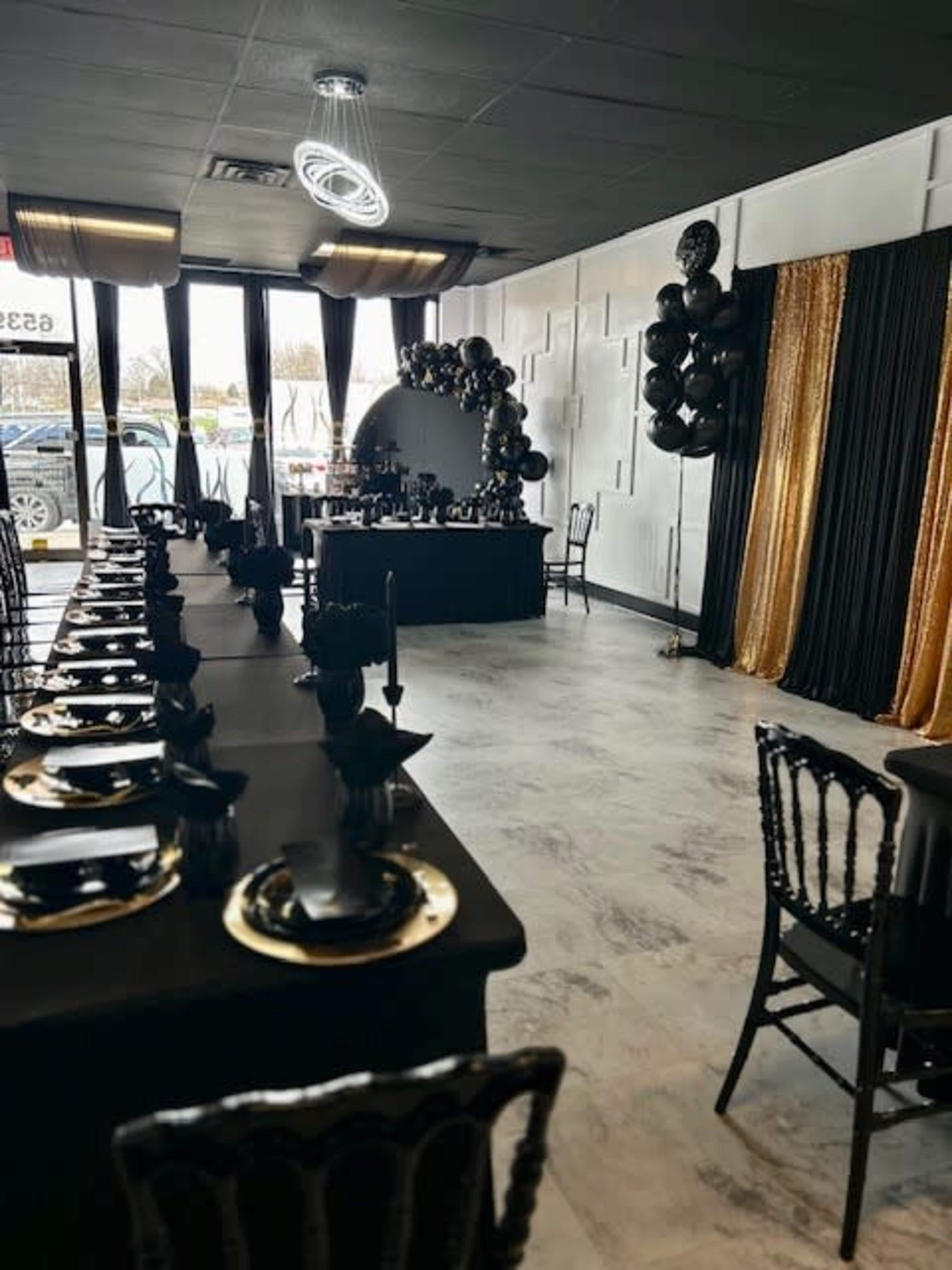 A banquet room is set up with long tables covered in black tablecloths, adorned with gold and black decorations, and a backdrop of black and gold balloons.