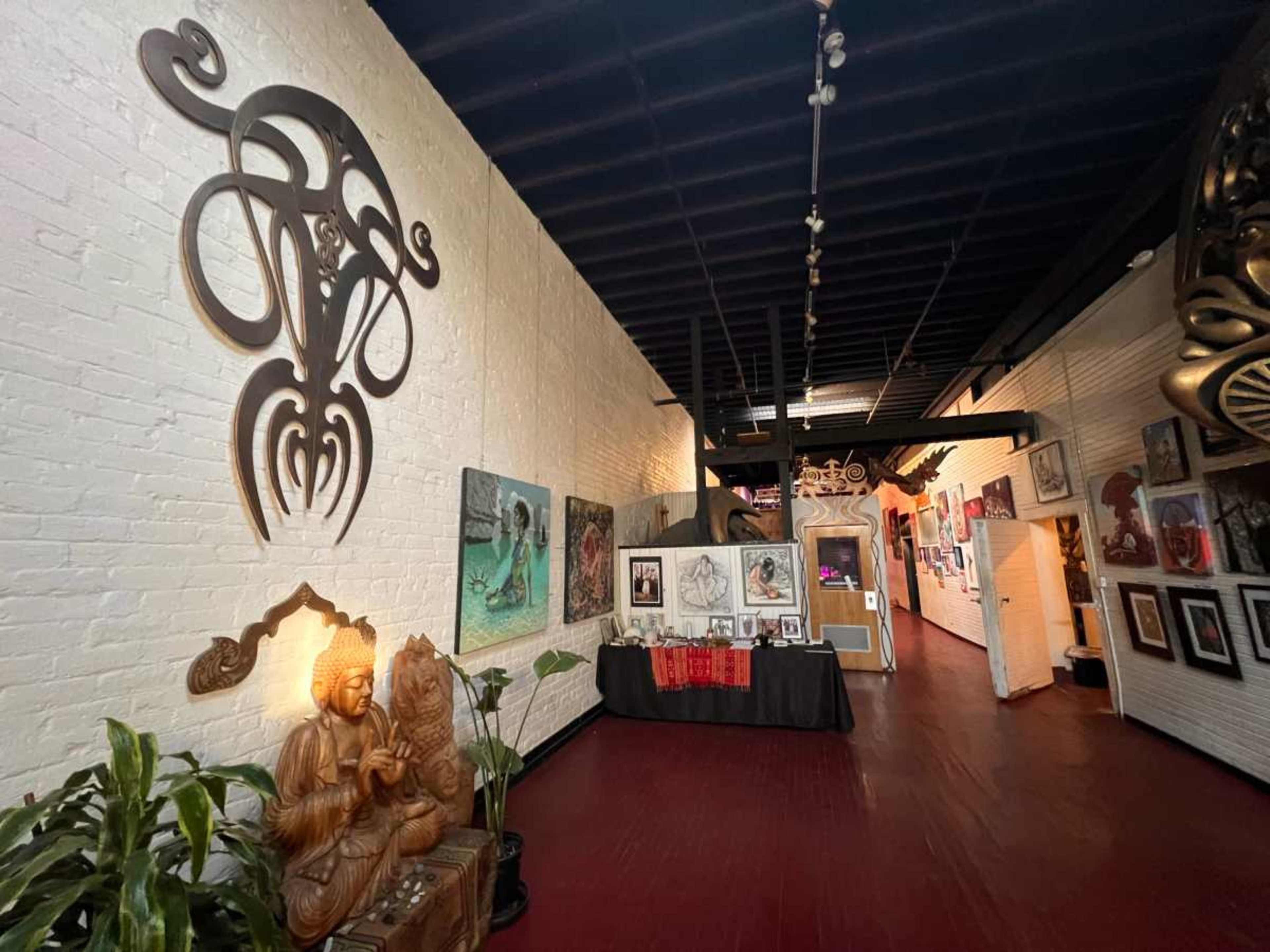 The image shows a long hallway in an art gallery with an ornate wall sculpture, various artworks displayed on the walls, and a table with framed pieces.