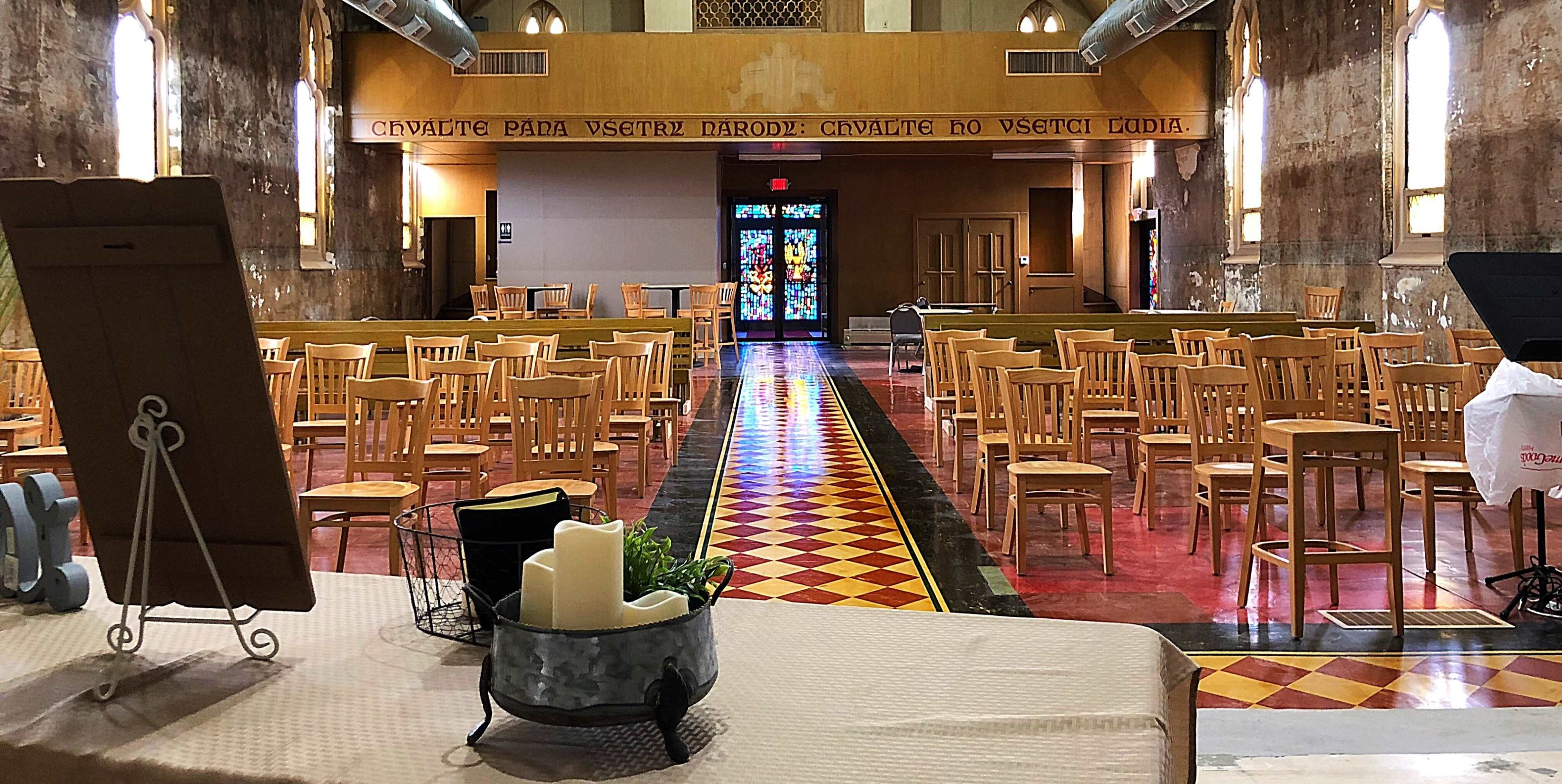 A spacious, empty hall with wooden chairs arranged in rows and a colorful stained glass window at the far end.