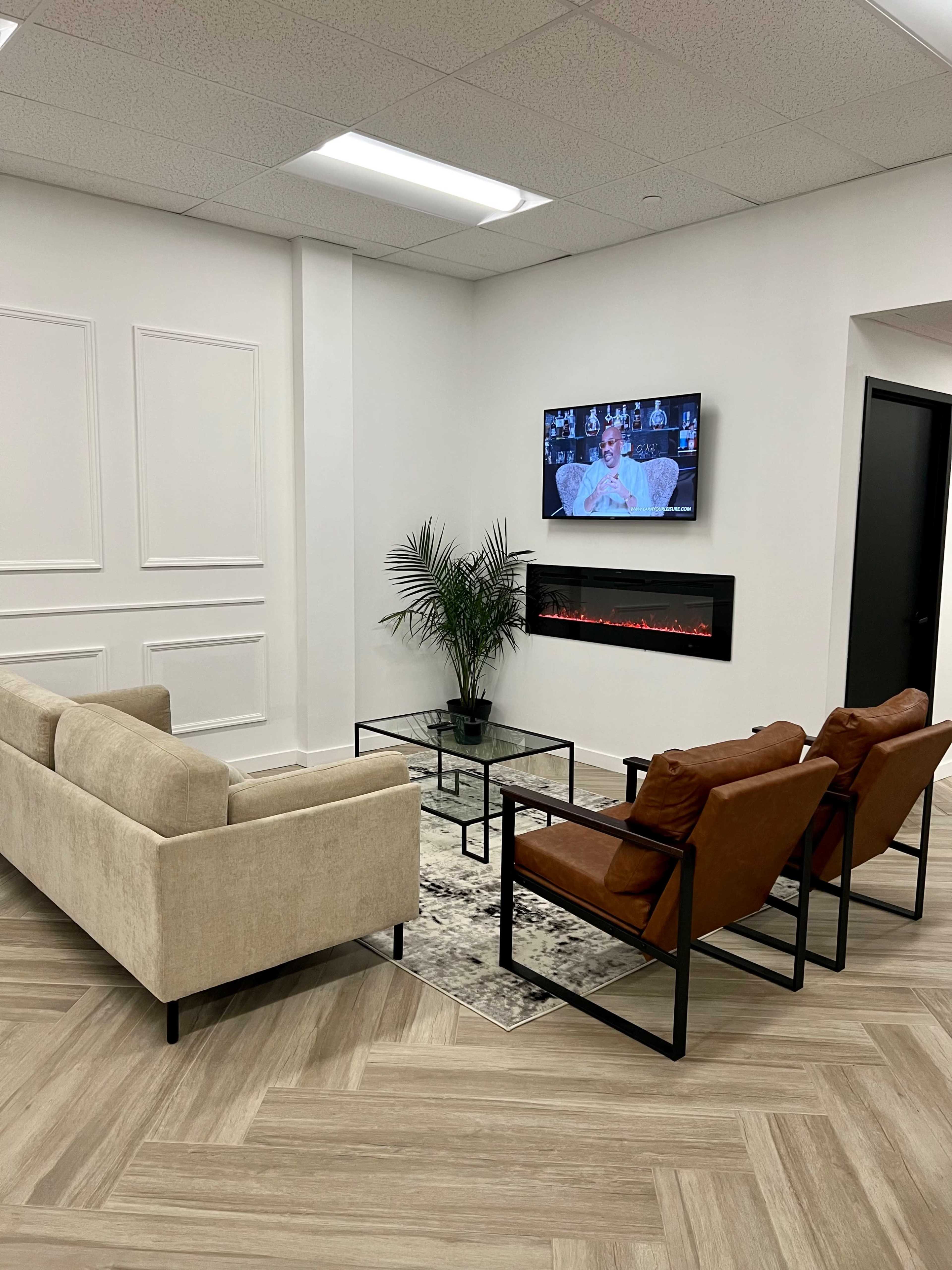 A modern waiting area featuring a beige sofa, two brown armchairs, a glass coffee table, an electric fireplace, and a wall-mounted television.