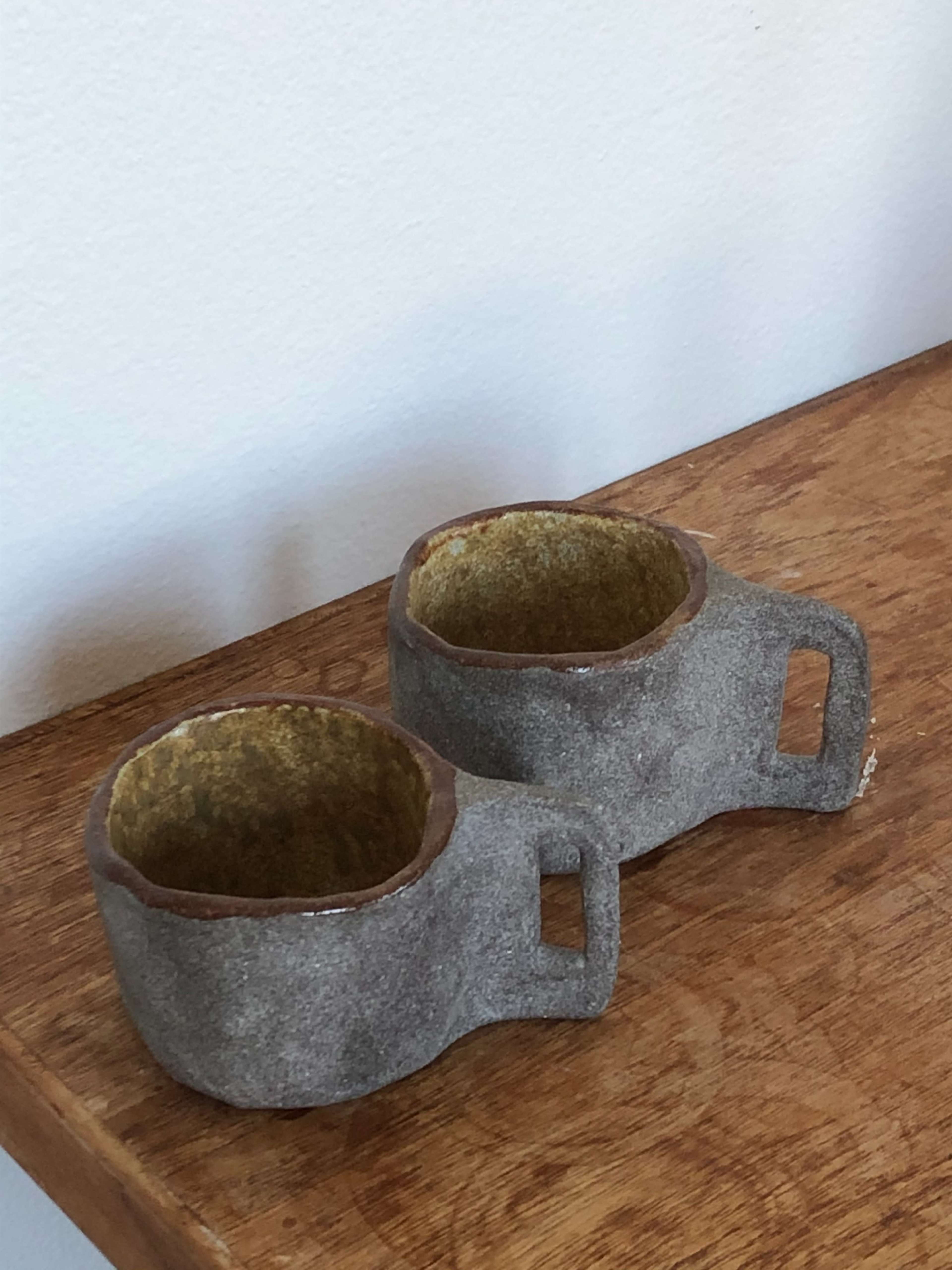 Two handmade ceramic cups with a textured gray exterior and yellow interior sit on a wooden surface.