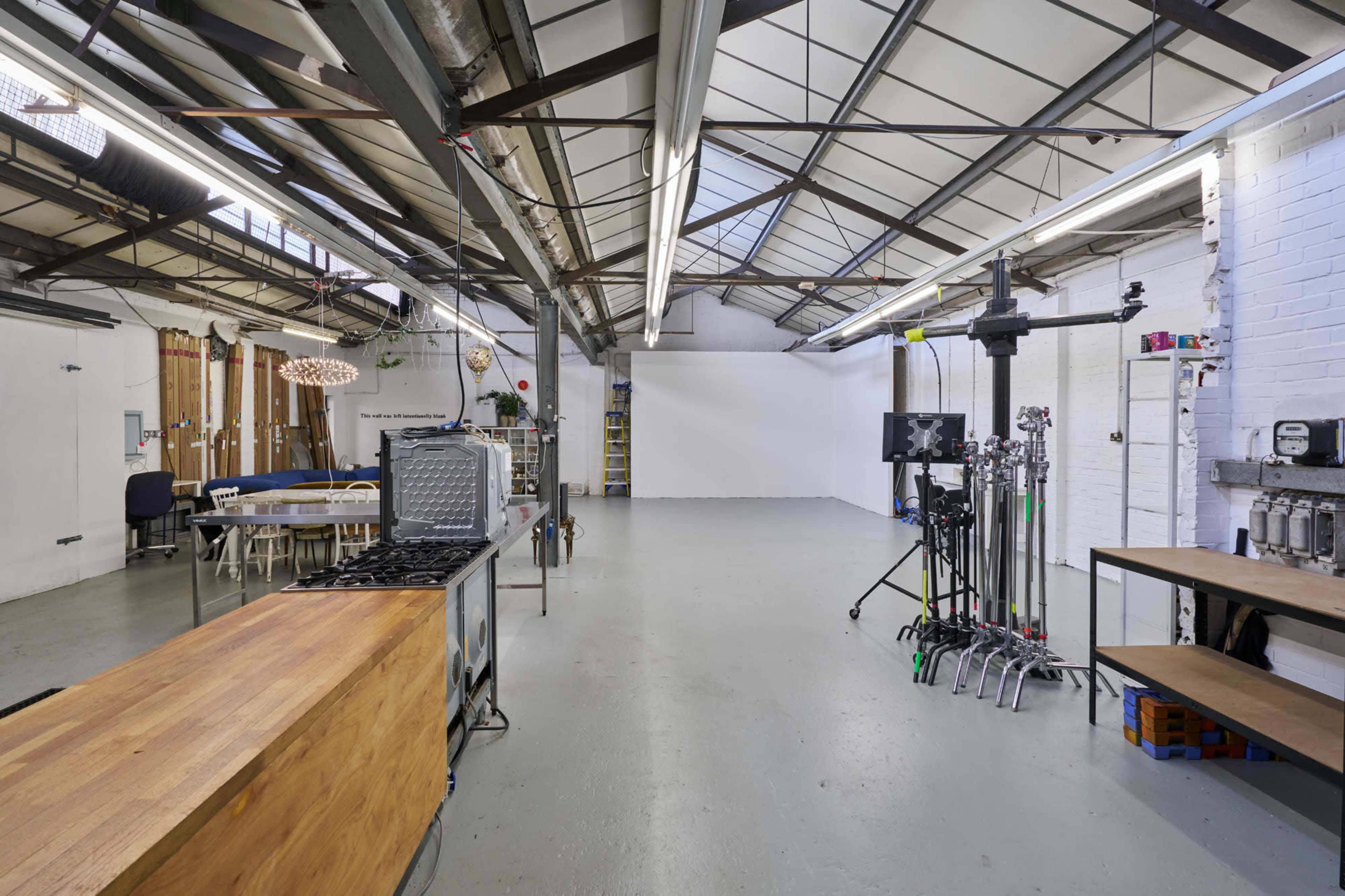 The image shows a spacious industrial-style studio with high ceilings, exposed beams, a kitchen area, various equipment on the floor, and a blank wall in the background.
