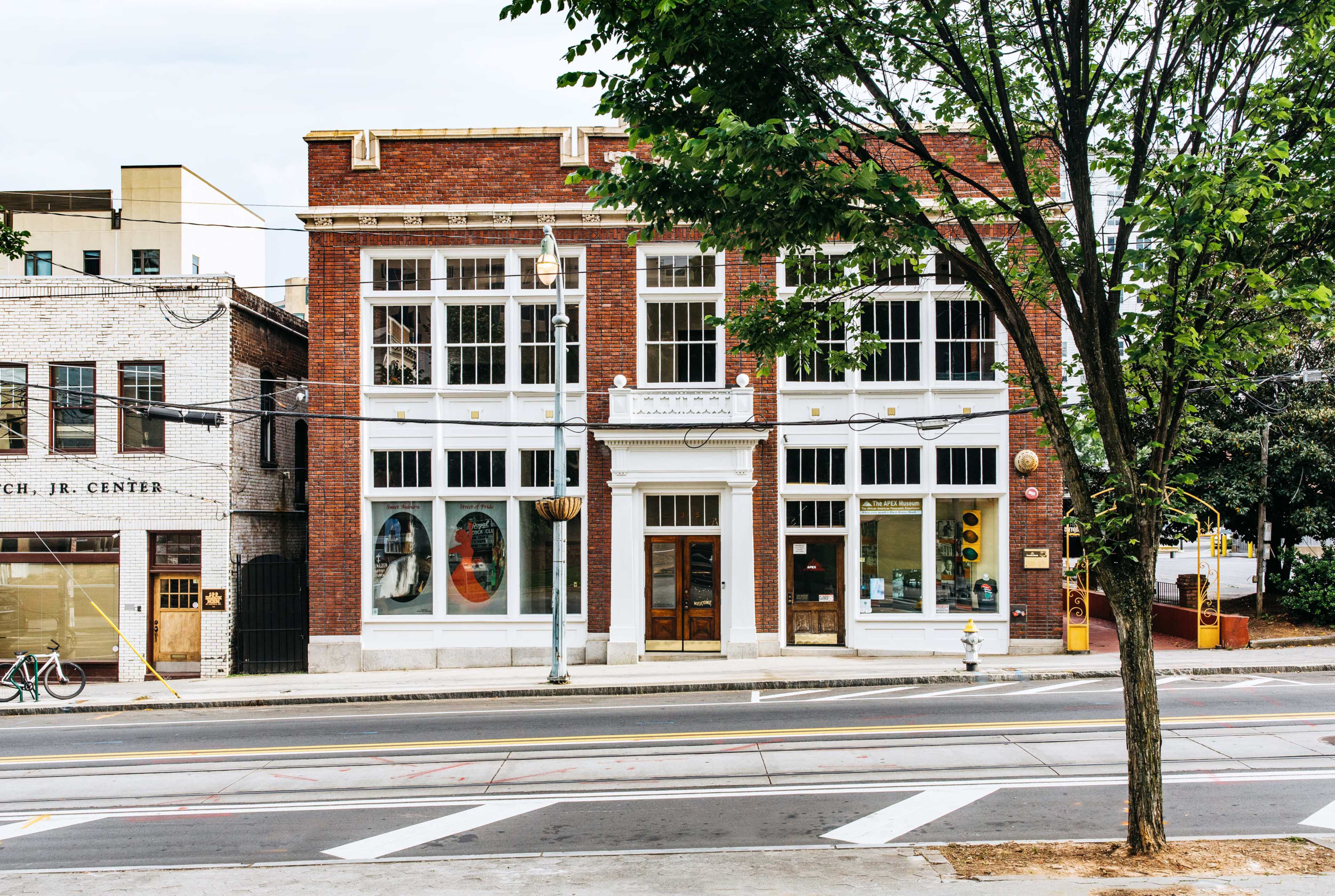 A red brick building with large windows and a white facade, situated along a street with a tree and bicycle nearby.