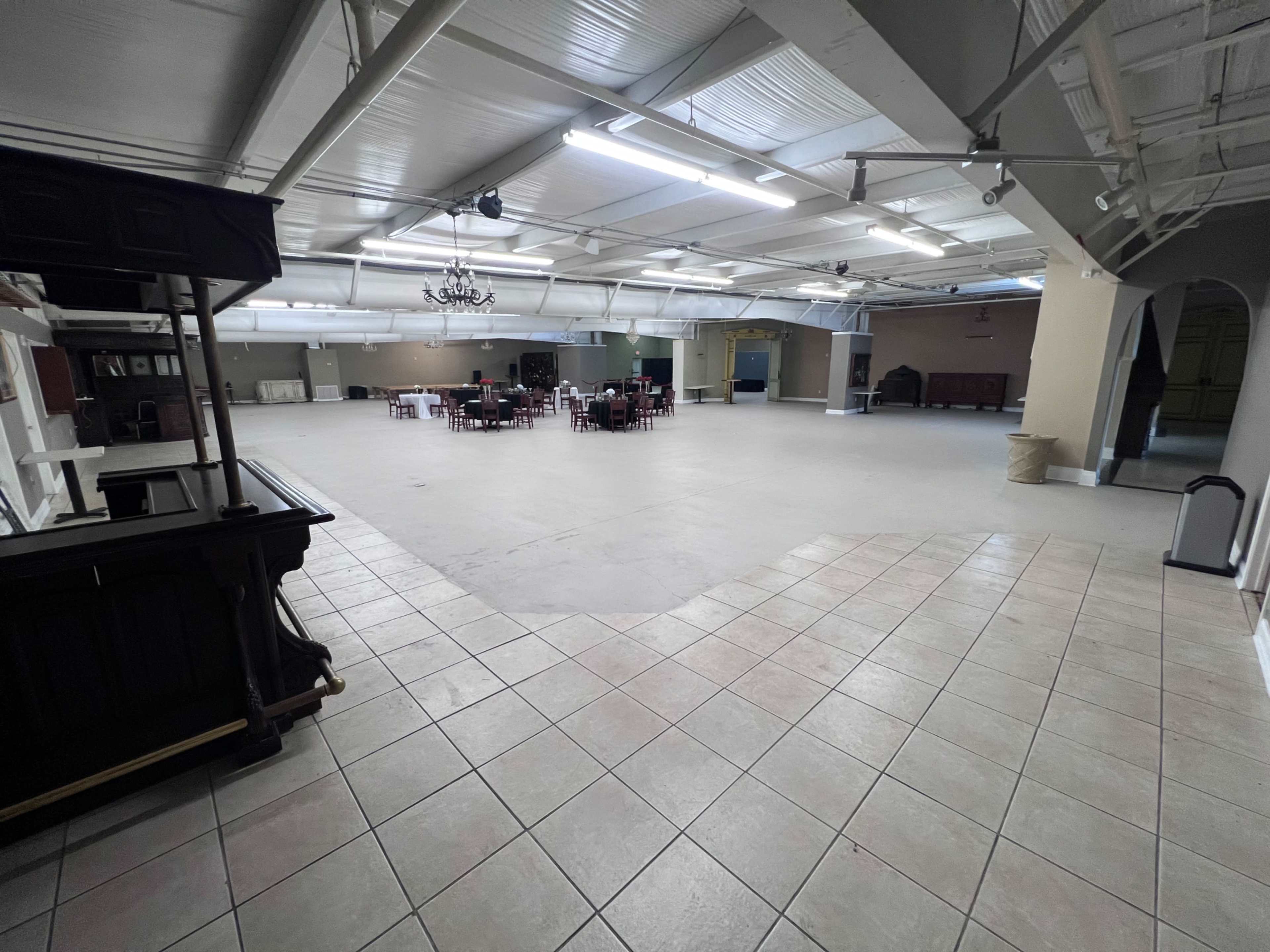 The image shows a large, empty hall with a tiled floor, sparse furnishings, and a few tables and chairs set up in the distance.