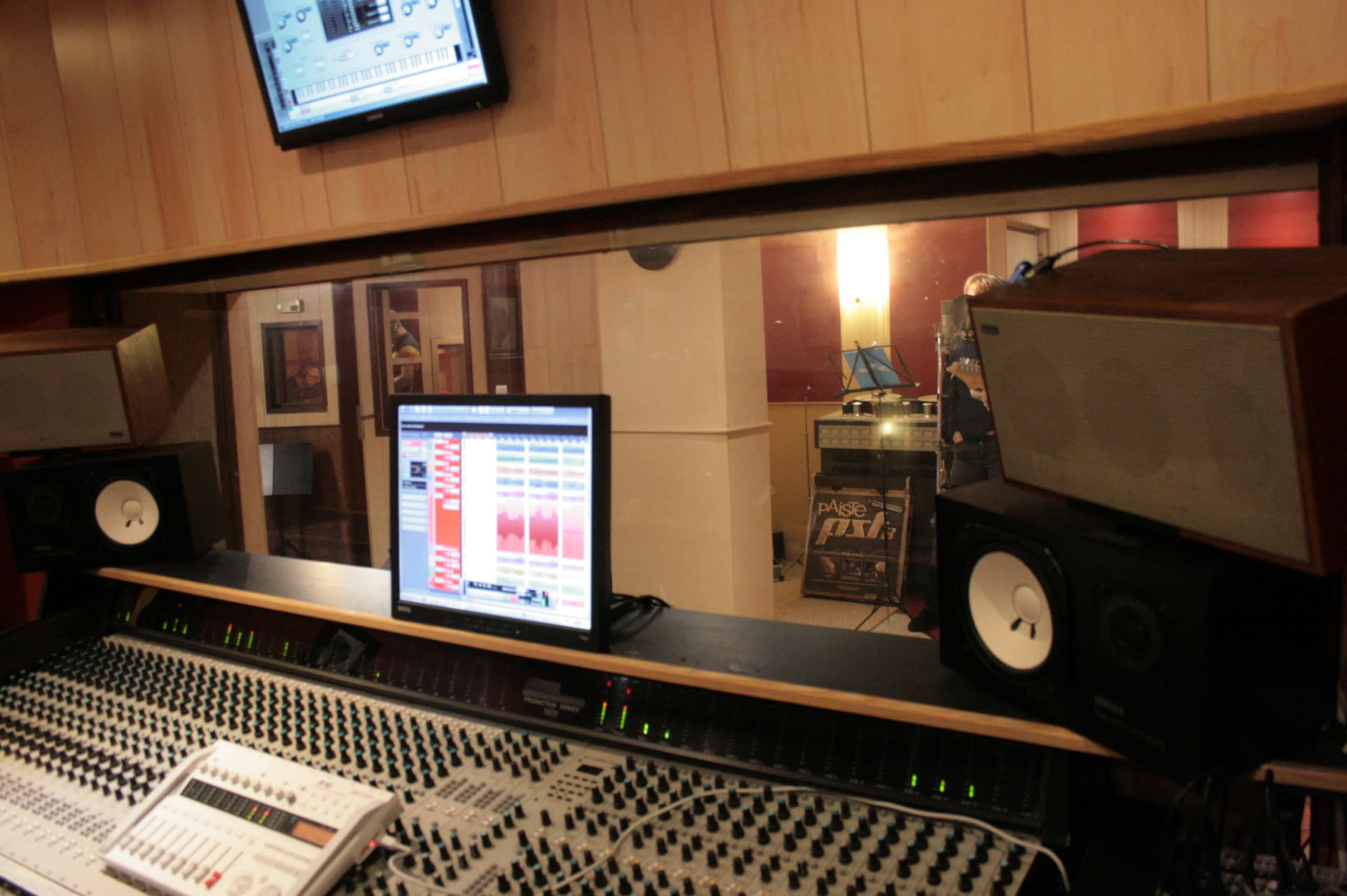 A recording studio control room features a large mixing console, computer monitor displaying audio tracks, and speakers, with a glimpse of the live room visible through a glass window.