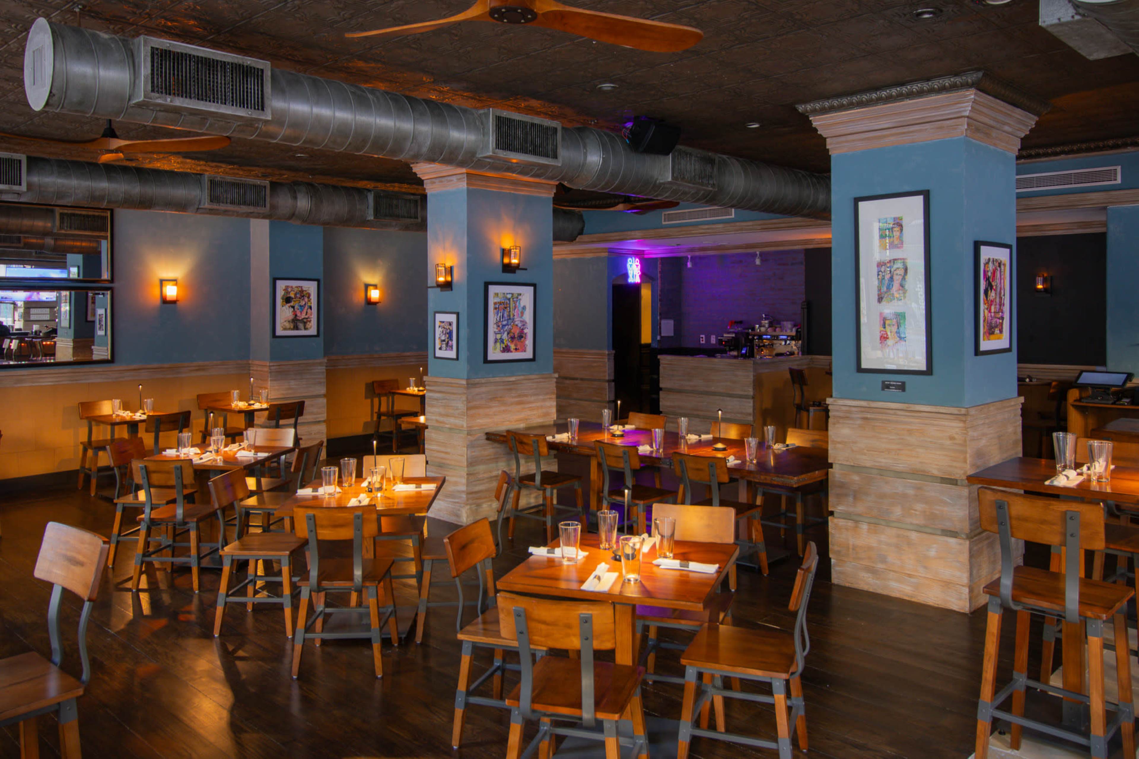 The image shows a restaurant interior with wooden tables and chairs arranged neatly, featuring a bar area in the background and a decorative ceiling.
