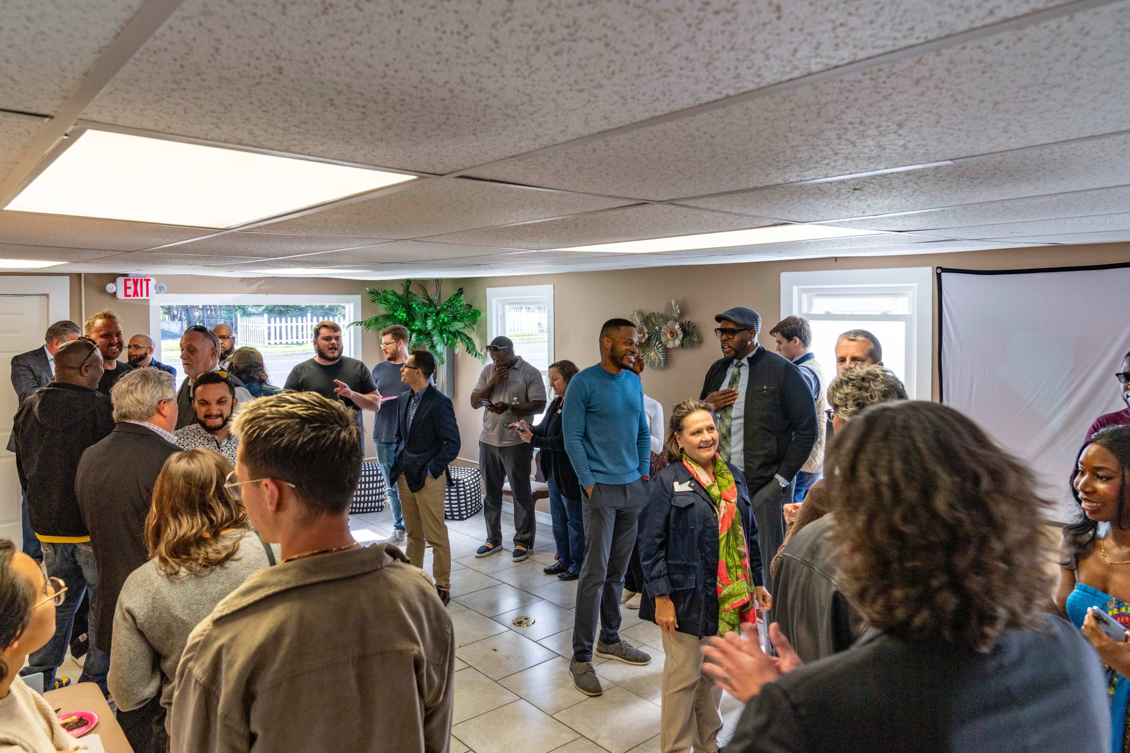 A diverse group of people socialize in a well-lit room with a tiled floor and windows.