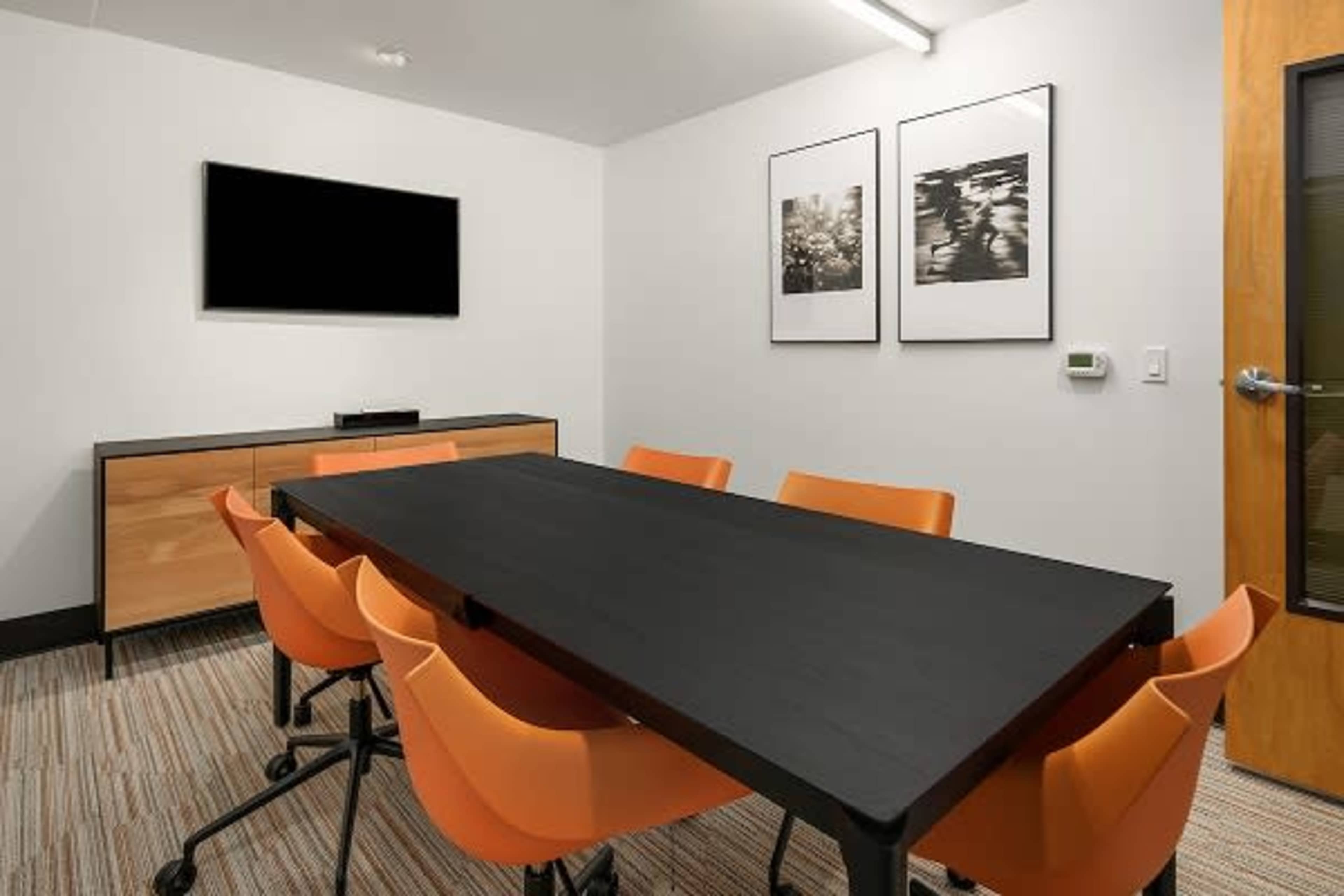 A modern conference room with a large black table surrounded by orange chairs, a television mounted on the wall, and two framed photographs.