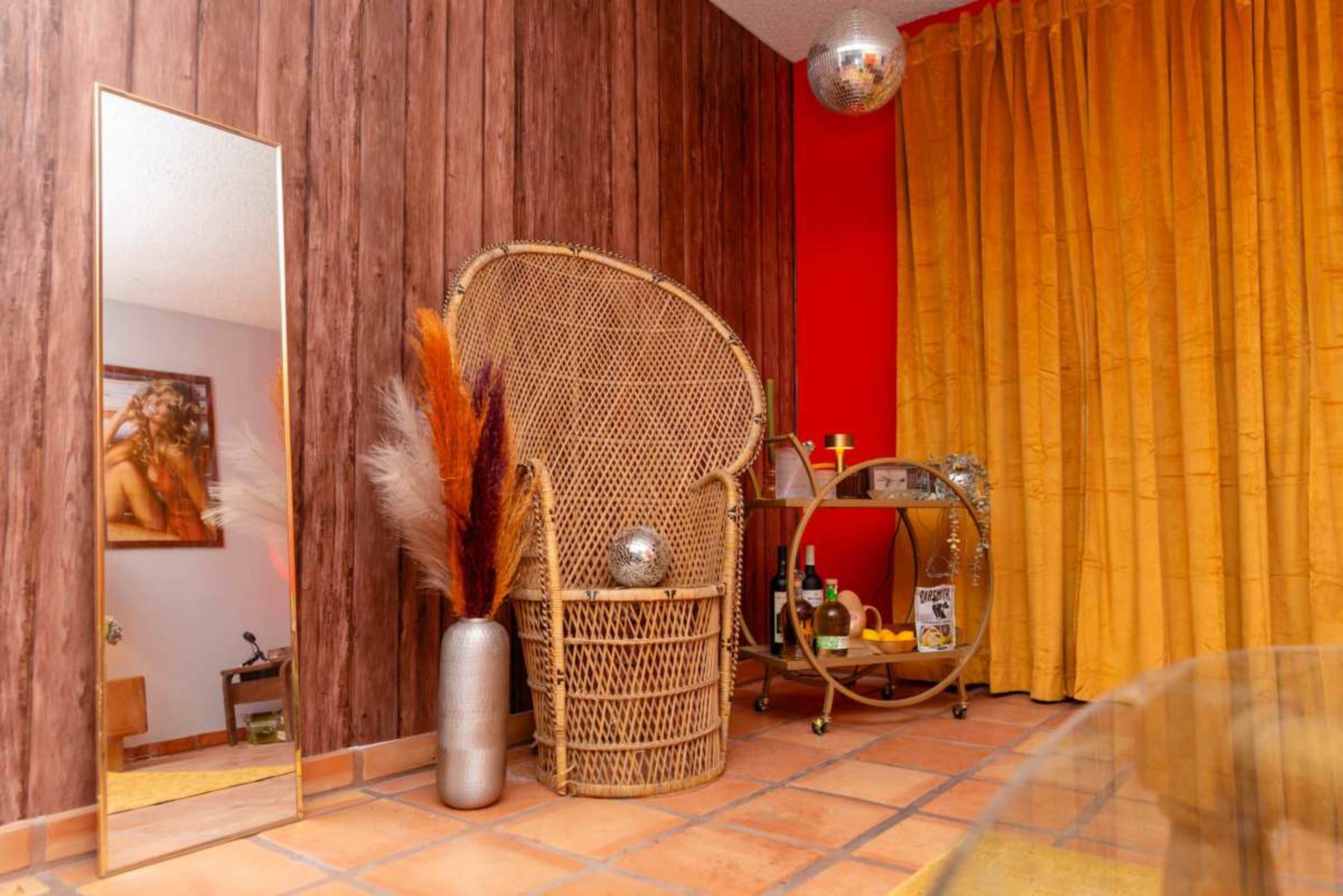 The image features a cozy corner of a room with a large wicker chair, a tall mirror, a decorative vase with dried plants, and a round bar cart adorned with drinks and a disco ball.