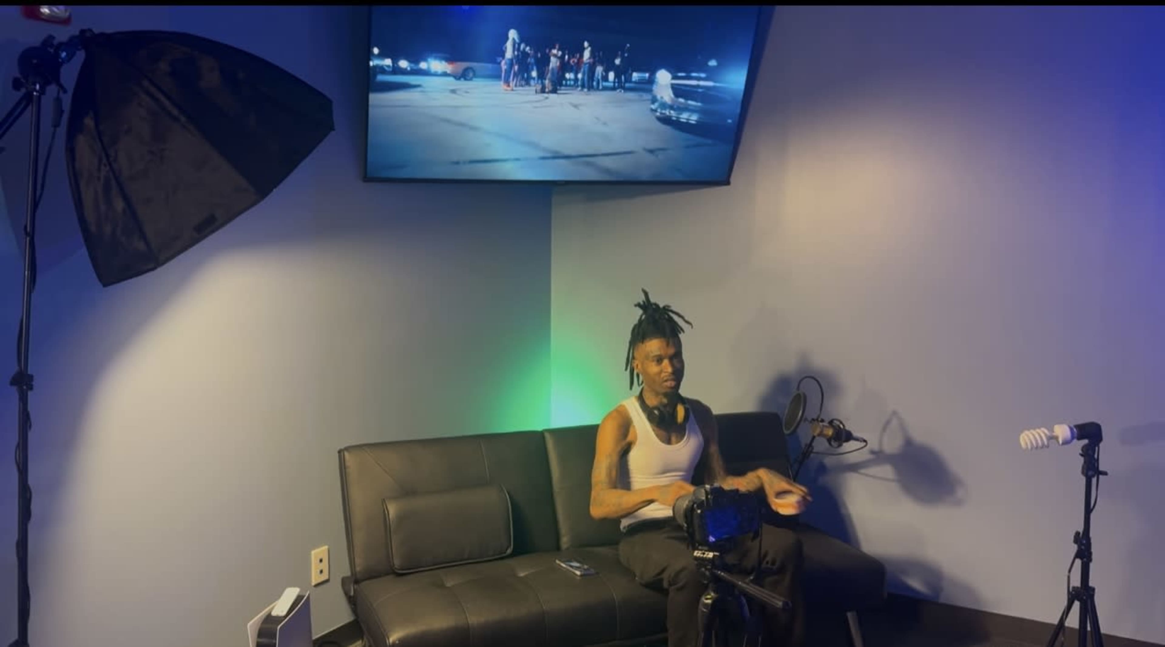 A person sits on a black couch in a room while watching a group of people on a screen in the background.