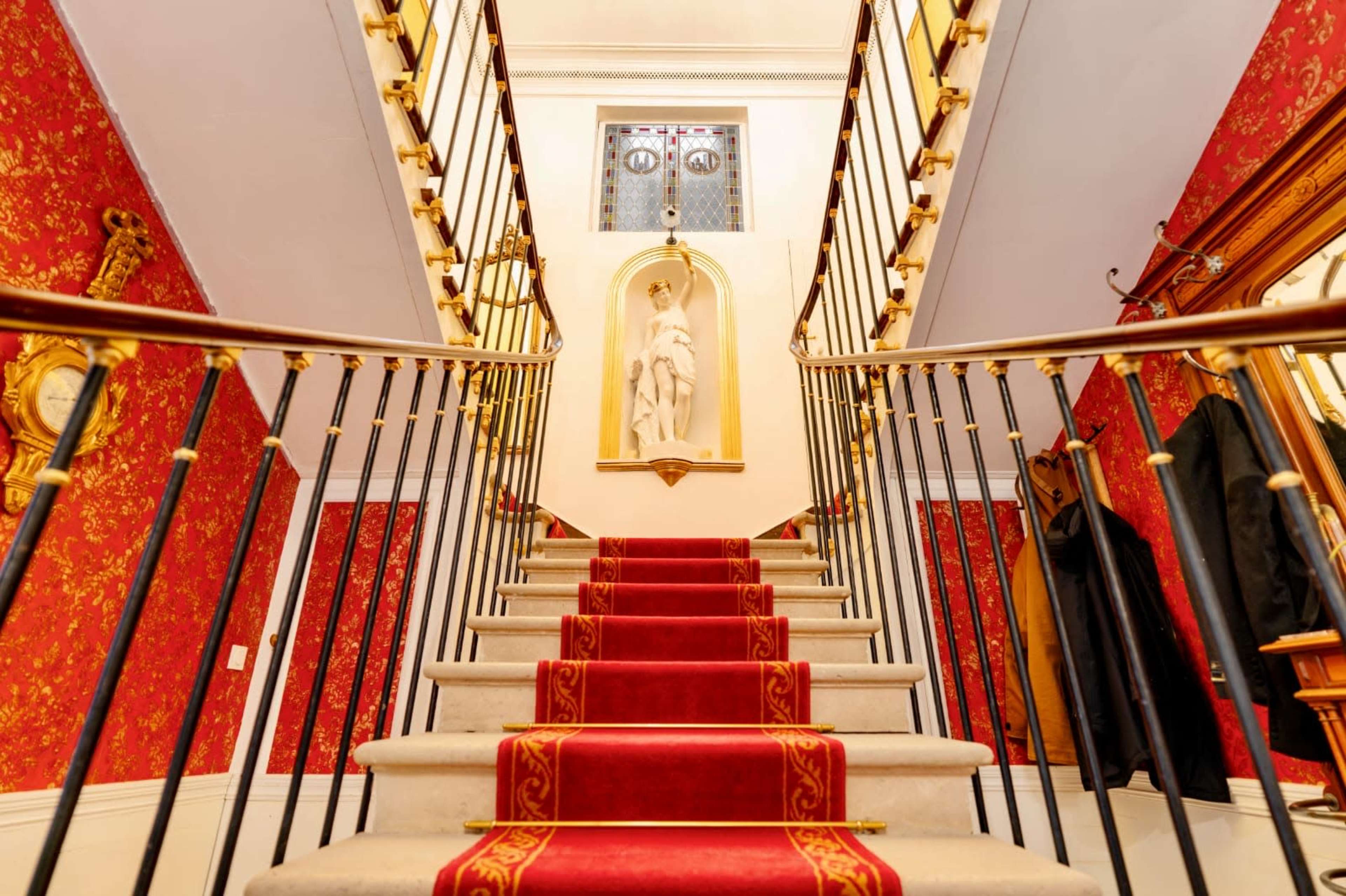 The image shows a staircase with red carpet leading up, flanked by ornate railings and decorated walls featuring red wallpaper.