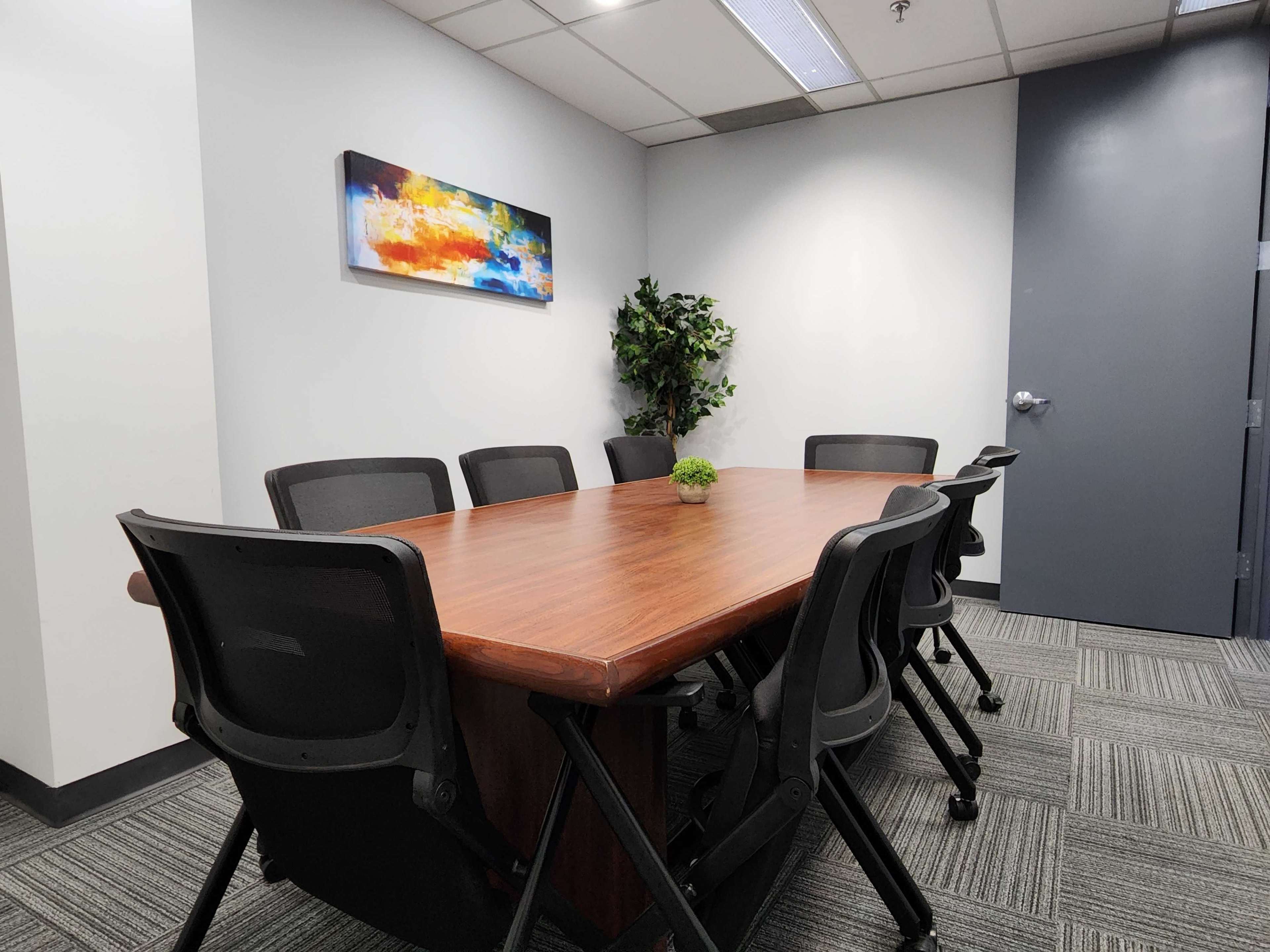 A conference room features a wooden table surrounded by black ergonomic chairs, with a small plant centerpiece and a colorful painting on the wall.