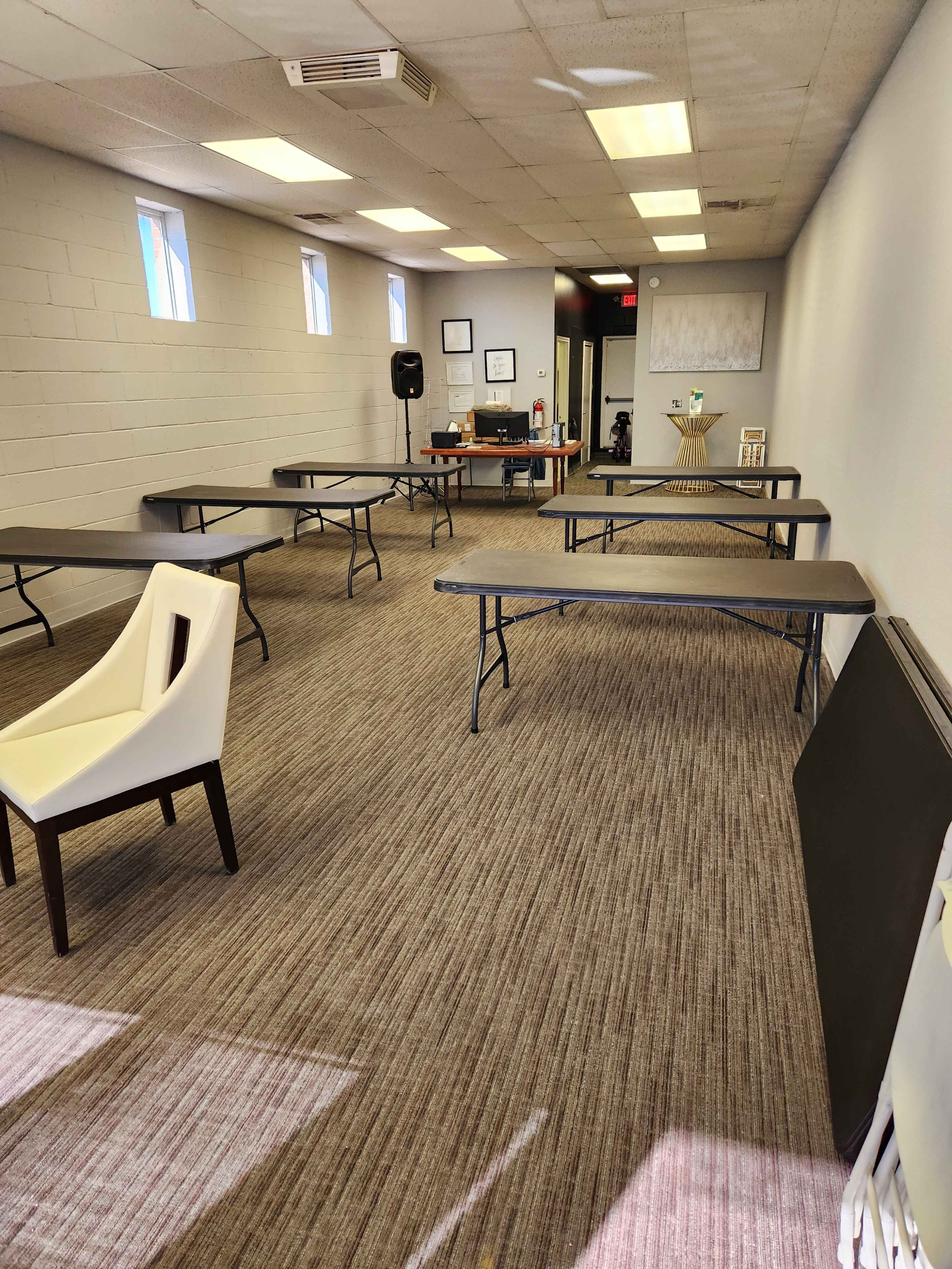 A sparse room with several folding tables arranged in a grid and a chair in the foreground, with windows providing natural light.