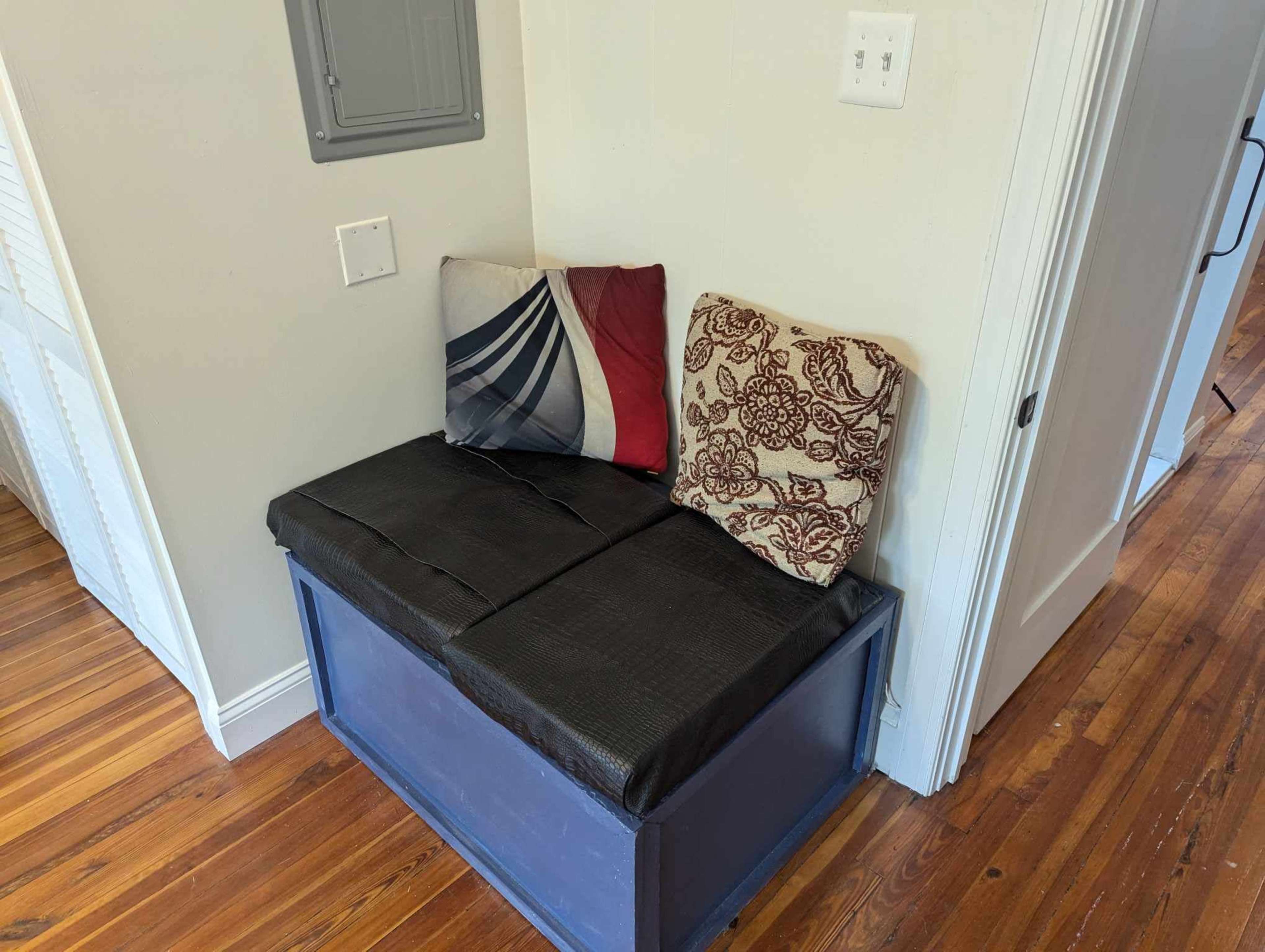 A small wooden bench with a black cushion and two decorative pillows is positioned against a wall in a room with wooden flooring.